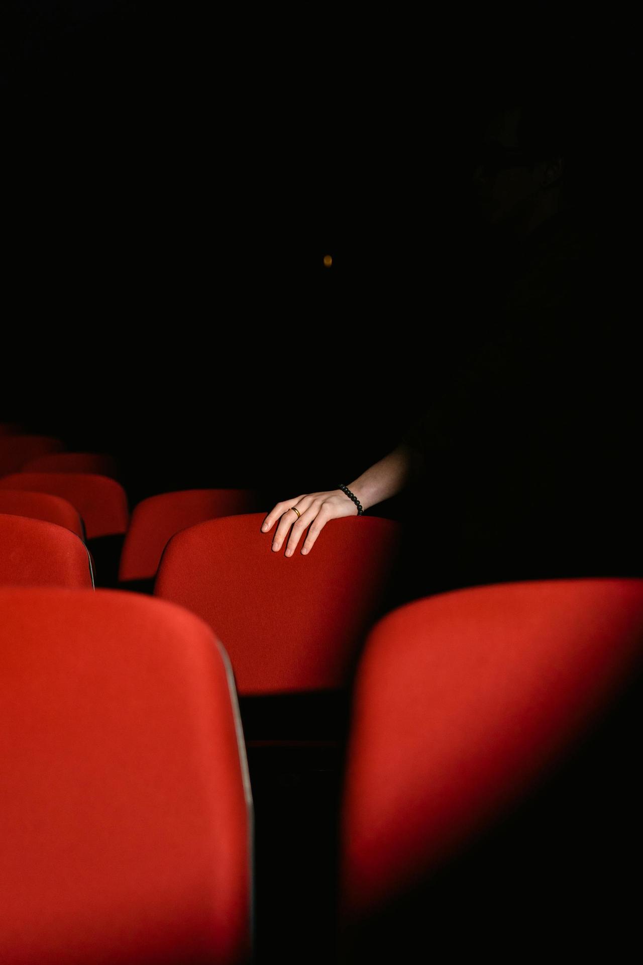 A hand rests on a red theater seat in dim lighting, creating a mysterious ambiance.