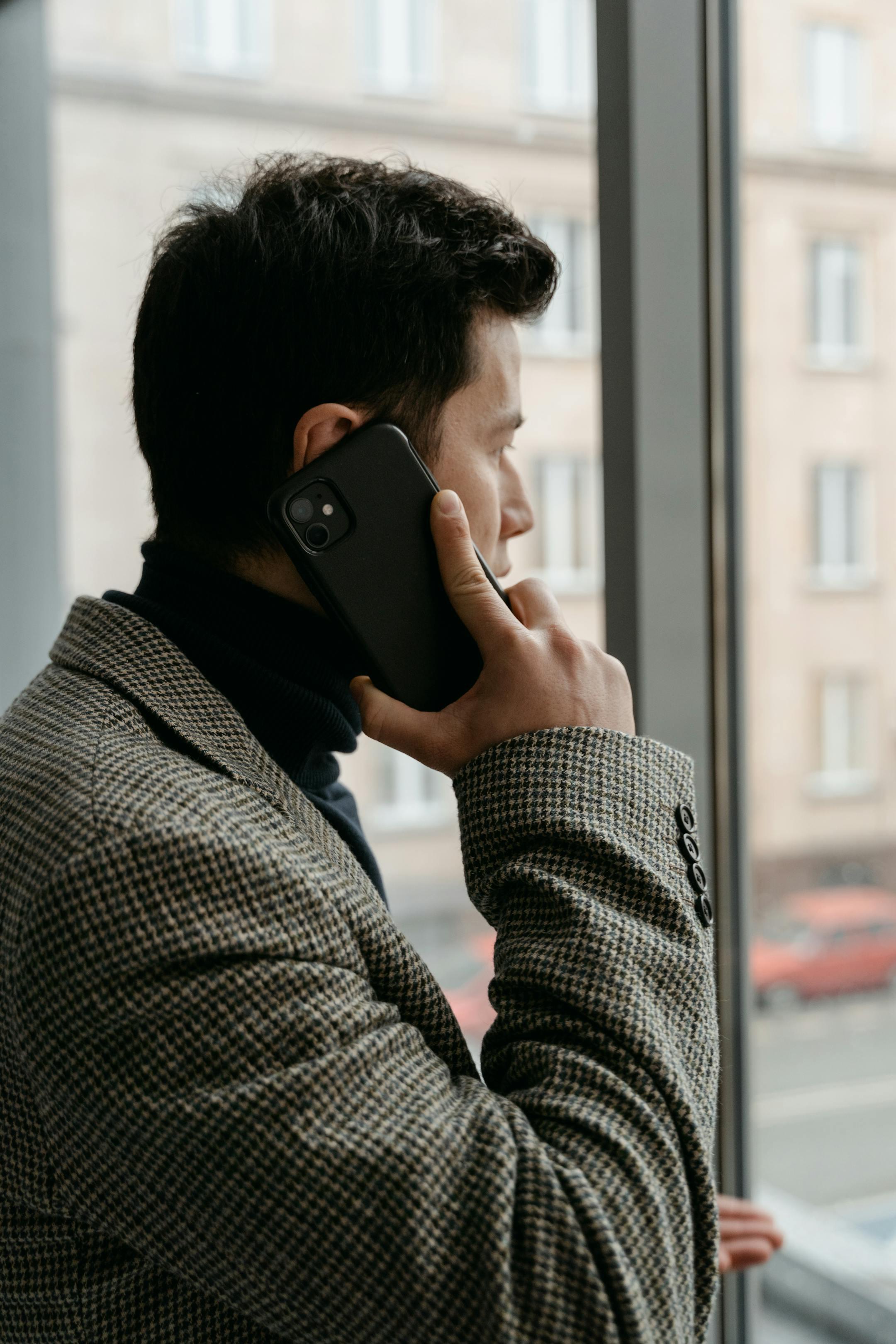 Man in a checkered blazer using a smartphone while gazing out a window.