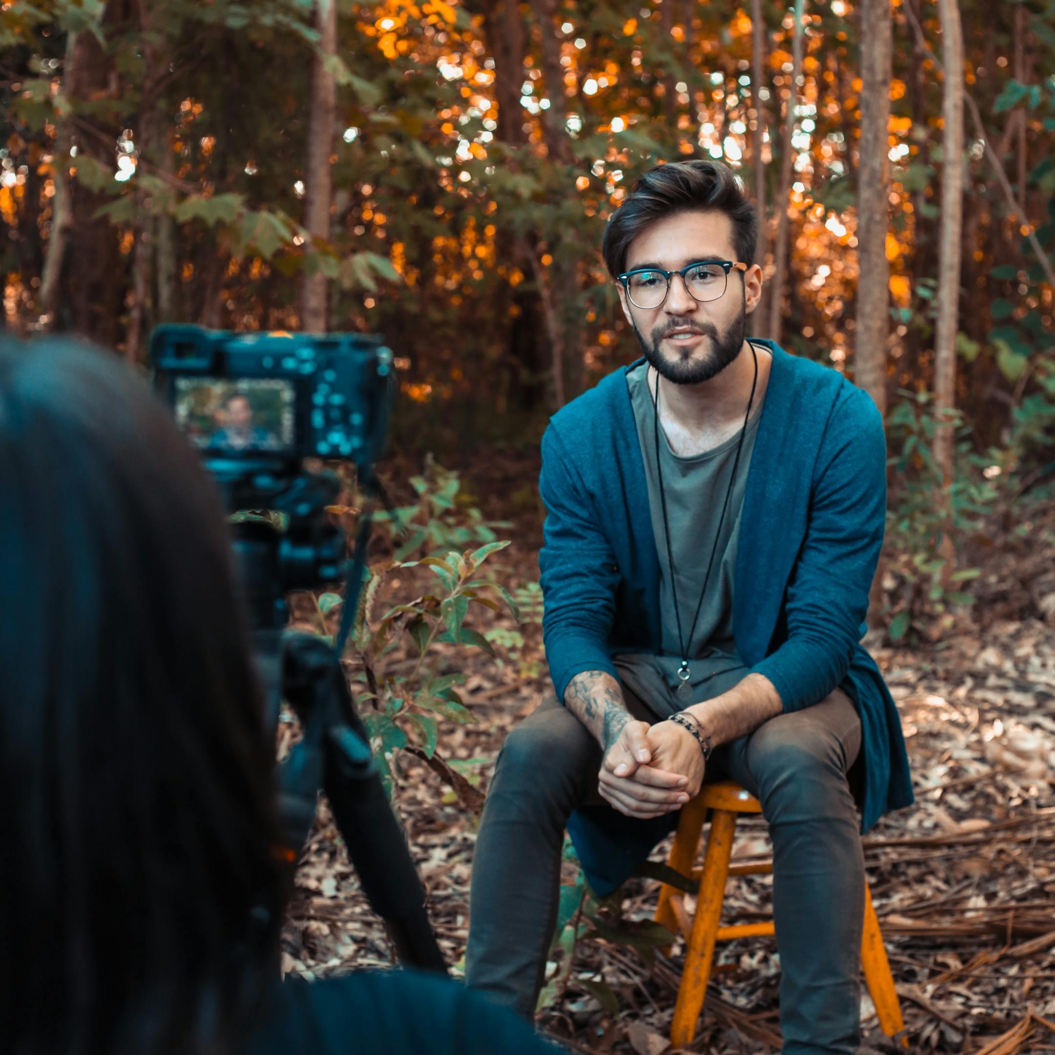 A young man being filmed outdoors in a forest, wearing casual attire and eyeglasses.
