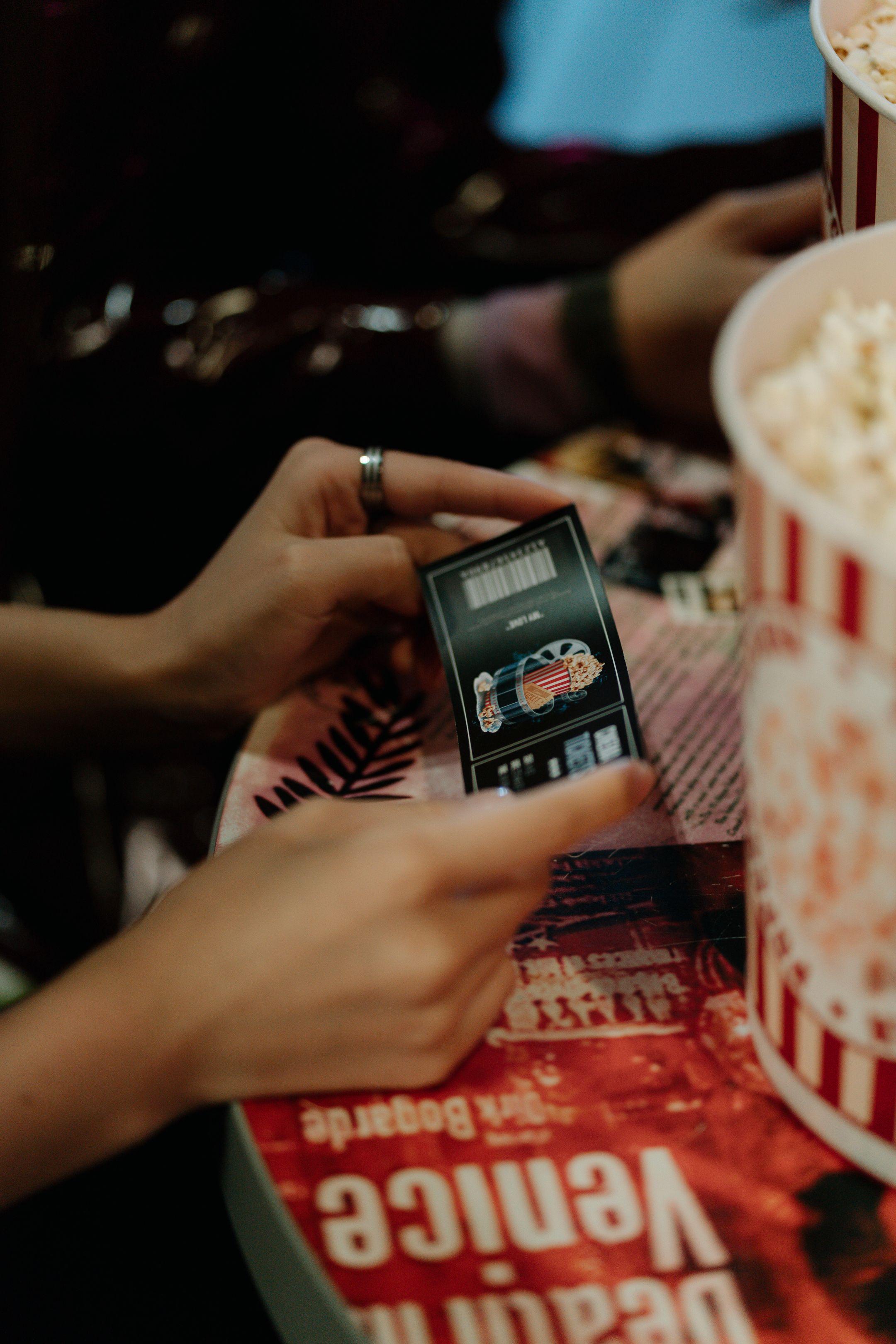 Close-up of hands holding a movie ticket with popcorn on a table.