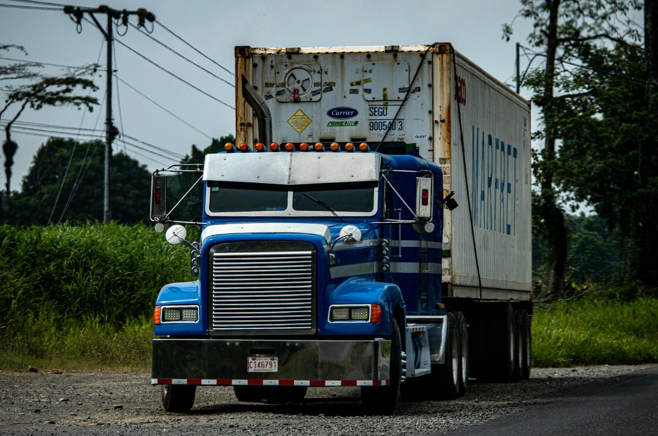 A close frontal view of a white semi-truck with visible grime and graffiti, driving on a road with tropical vegetation in the background.