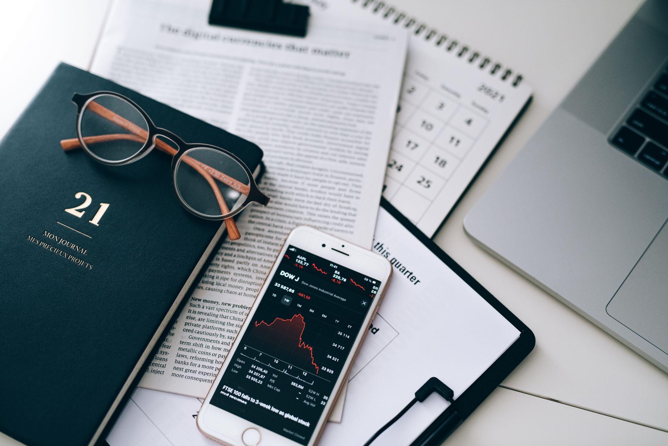 A stylish workspace featuring financial documents, eyeglasses, an iPhone displaying stock data, and a laptop.