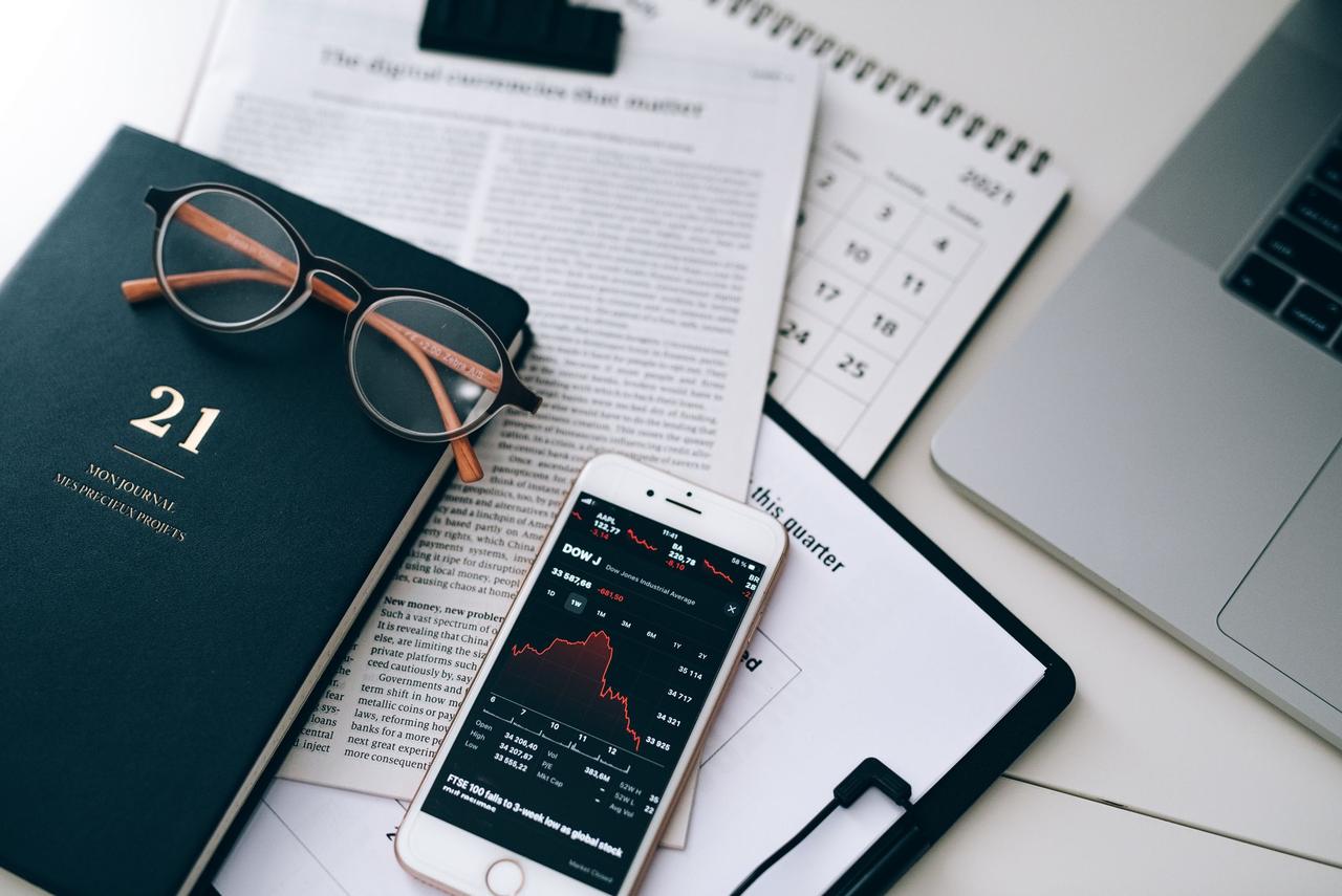A stylish workspace featuring financial documents, eyeglasses, an iPhone displaying stock data, and a laptop.