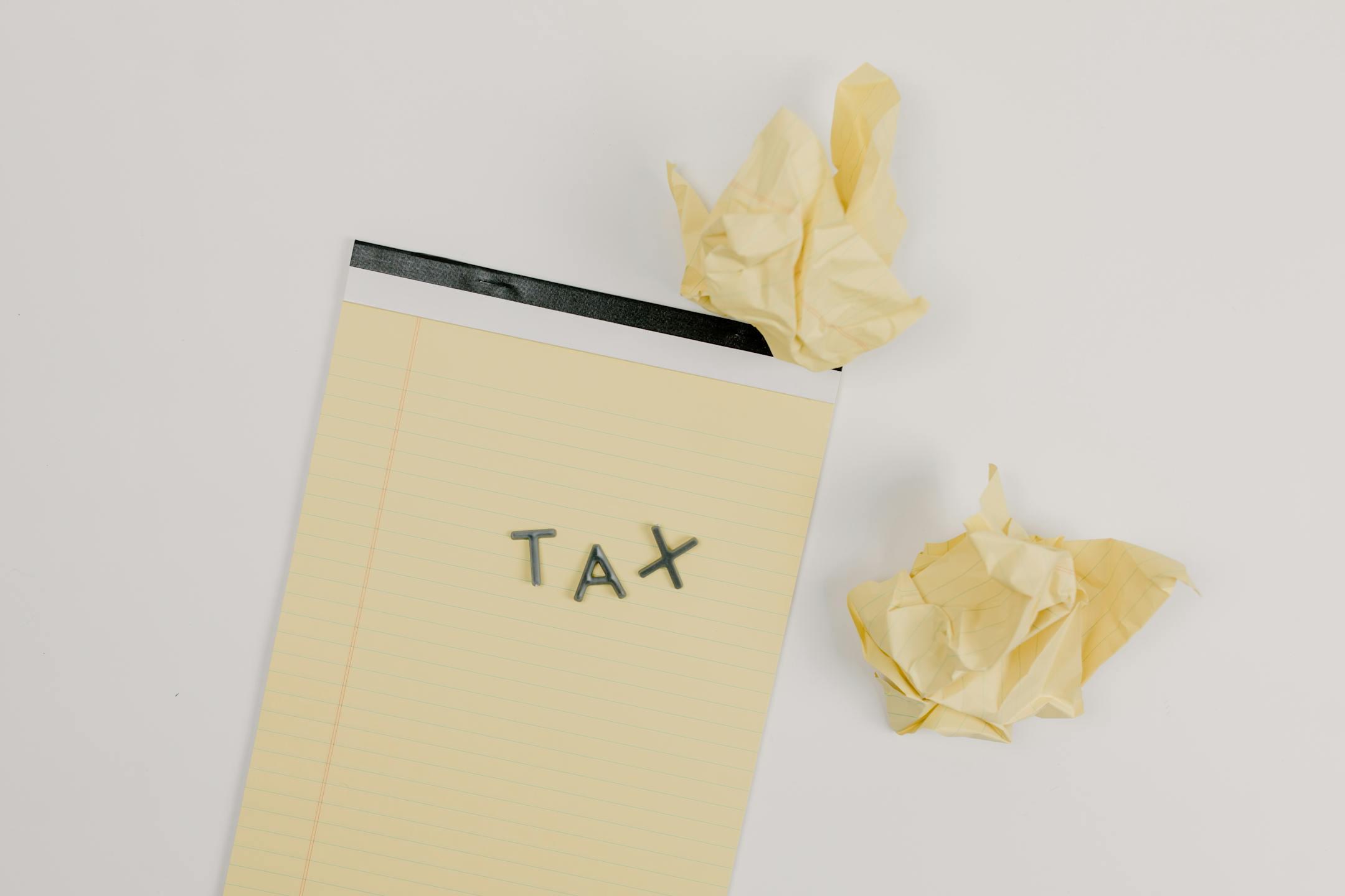 Flat lay of a notepad labeled 'tax' with crumpled paper on a white background, symbolizing financial stress.