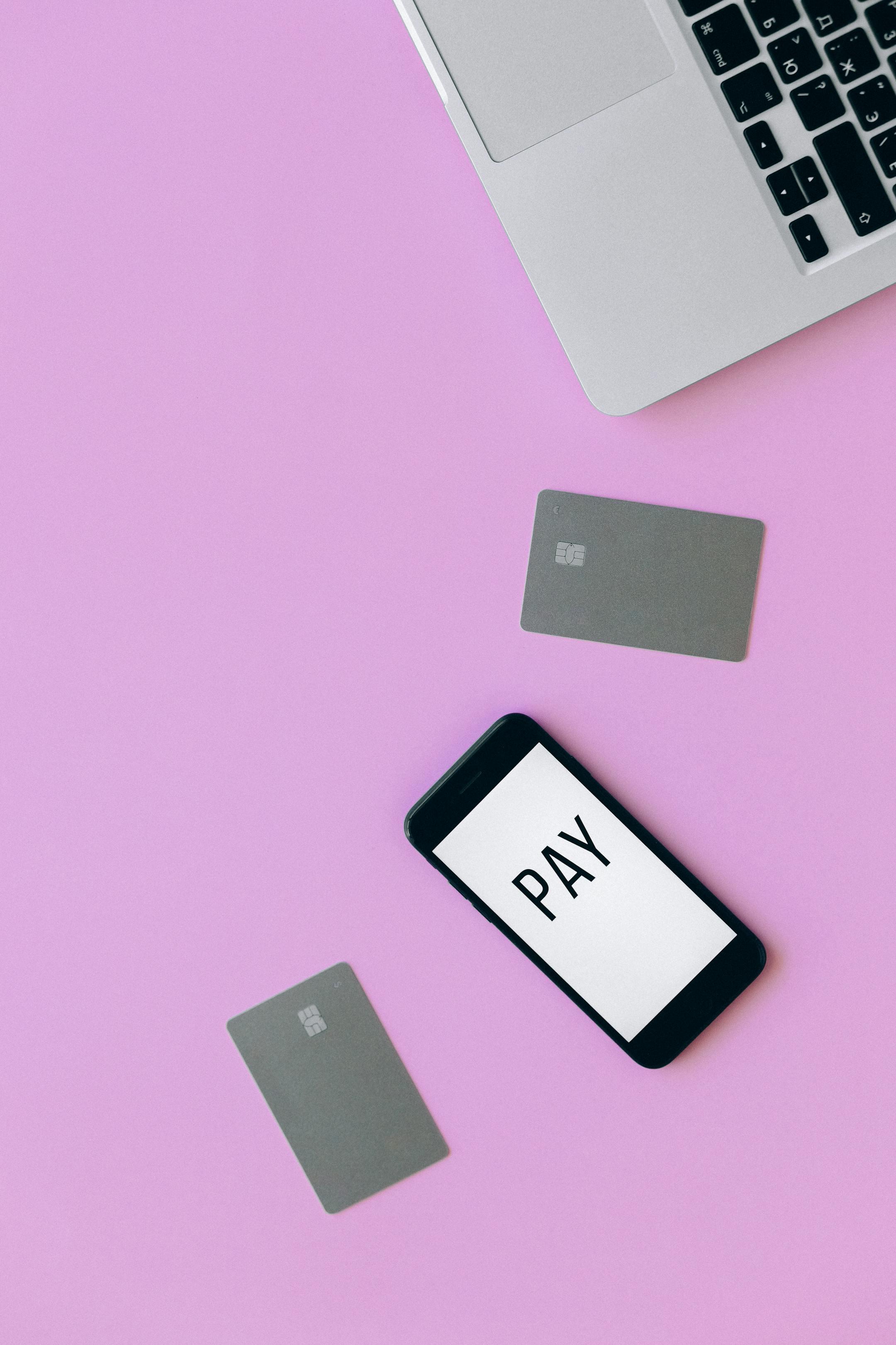 Flat lay of a laptop, smartphone displaying 'PAY', and credit cards on a pink surface representing online transaction.