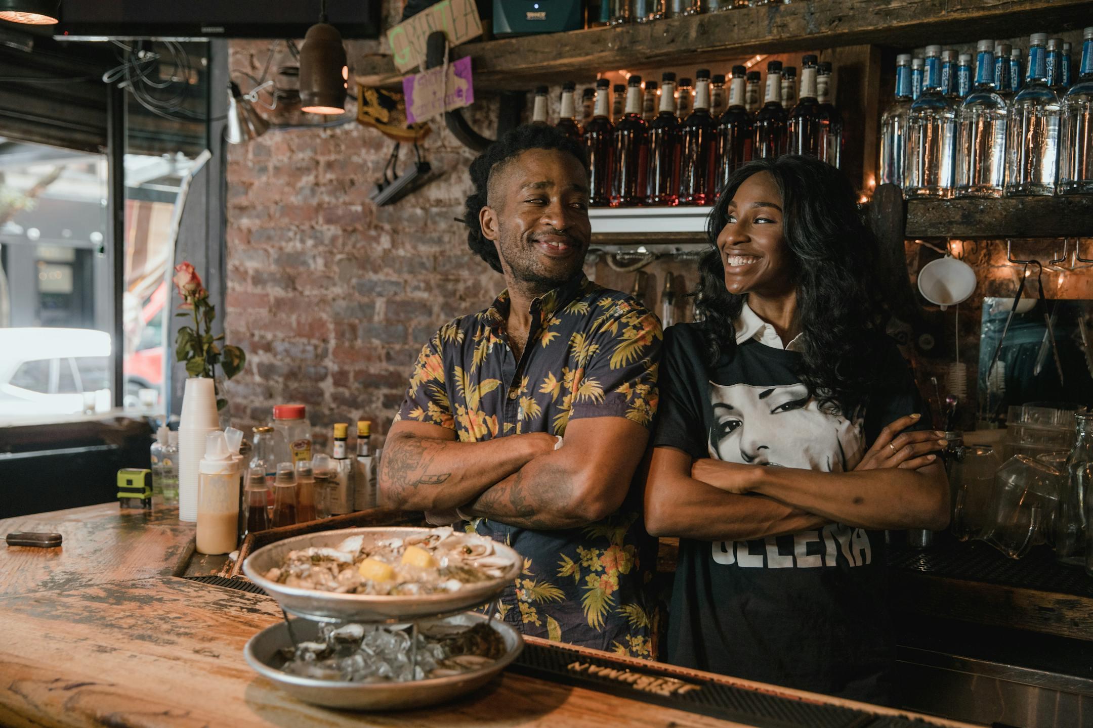 Two Black business owners smiling confidently behind a bar with oysters, embodying success and pride.