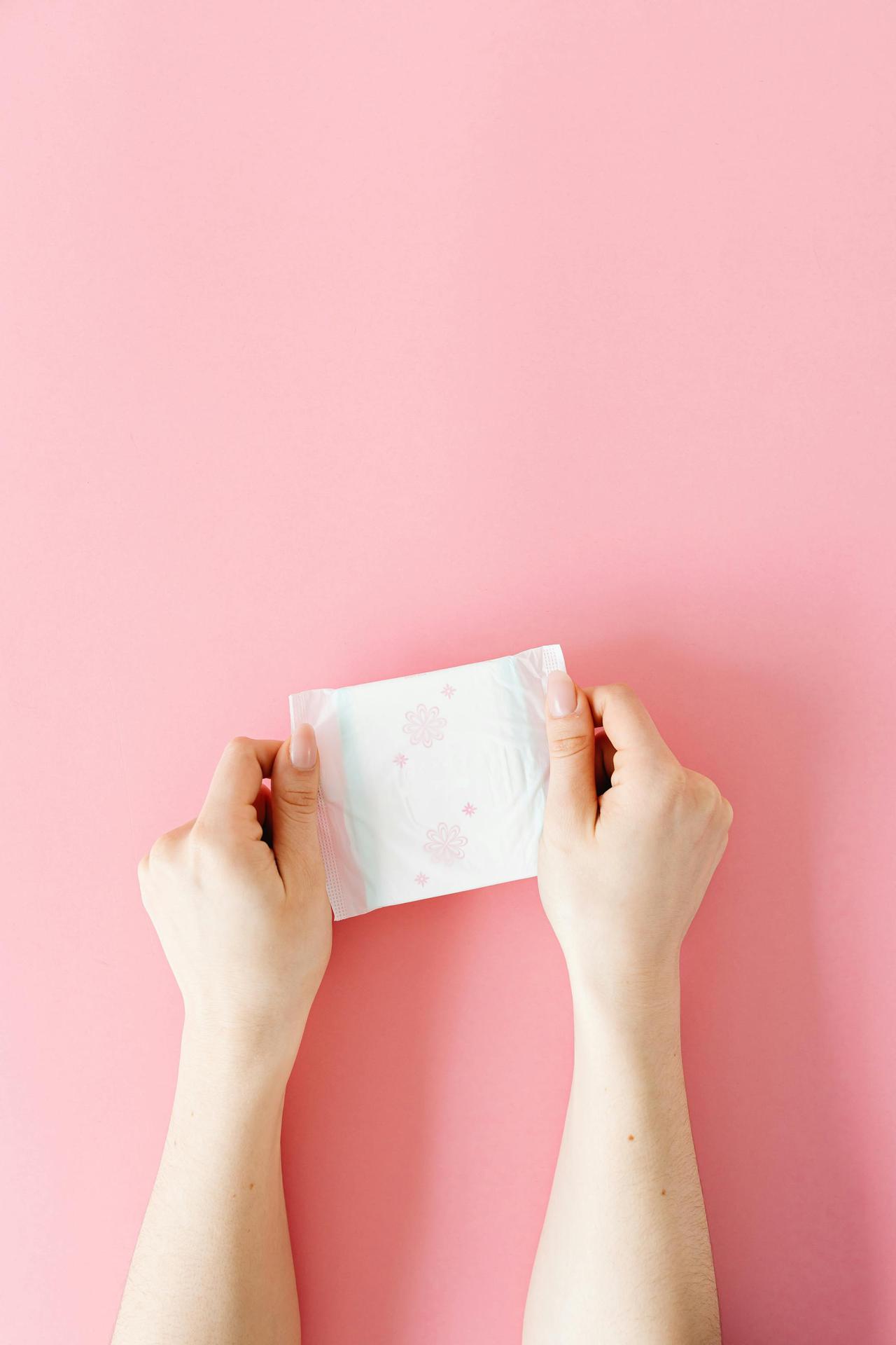 Minimalist photo of hands holding a sanitary pad on a pink background, symbolizing feminine hygiene.