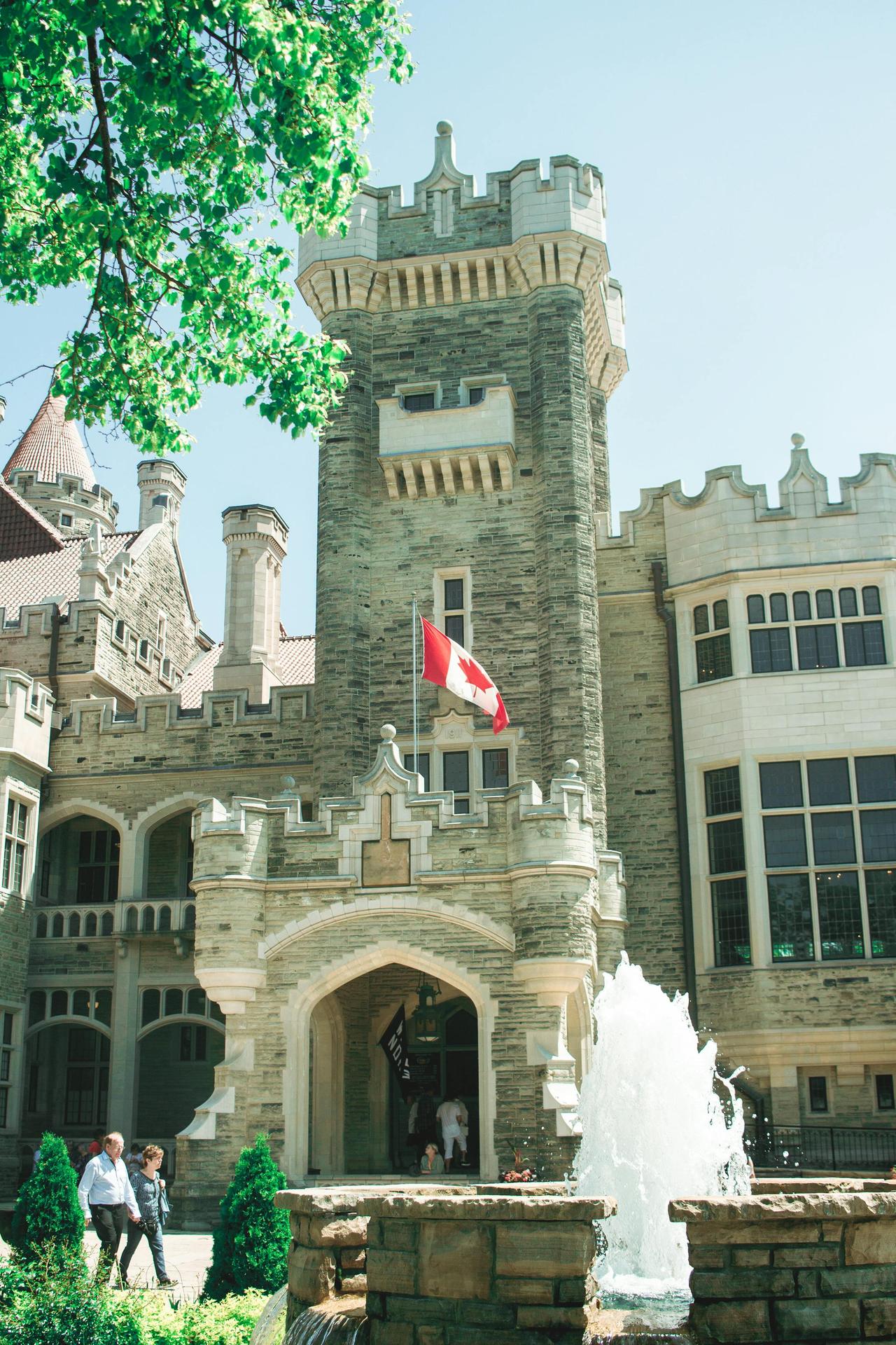 Historic Casa Loma castle with Canadian flag and fountain under blue sky.
