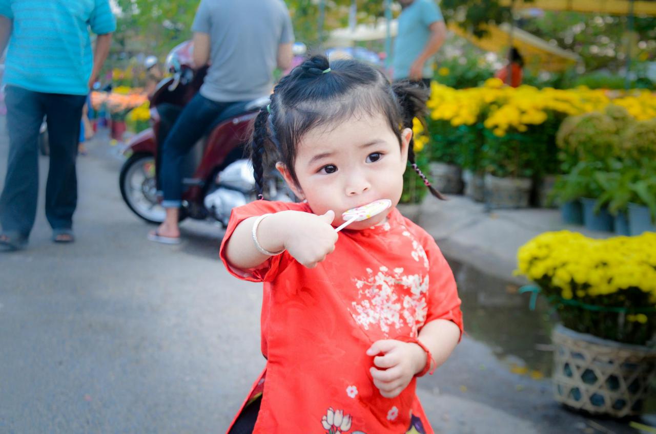 Cute toddler girl in red traditional dress eating a lollipop in a vibrant park setting.