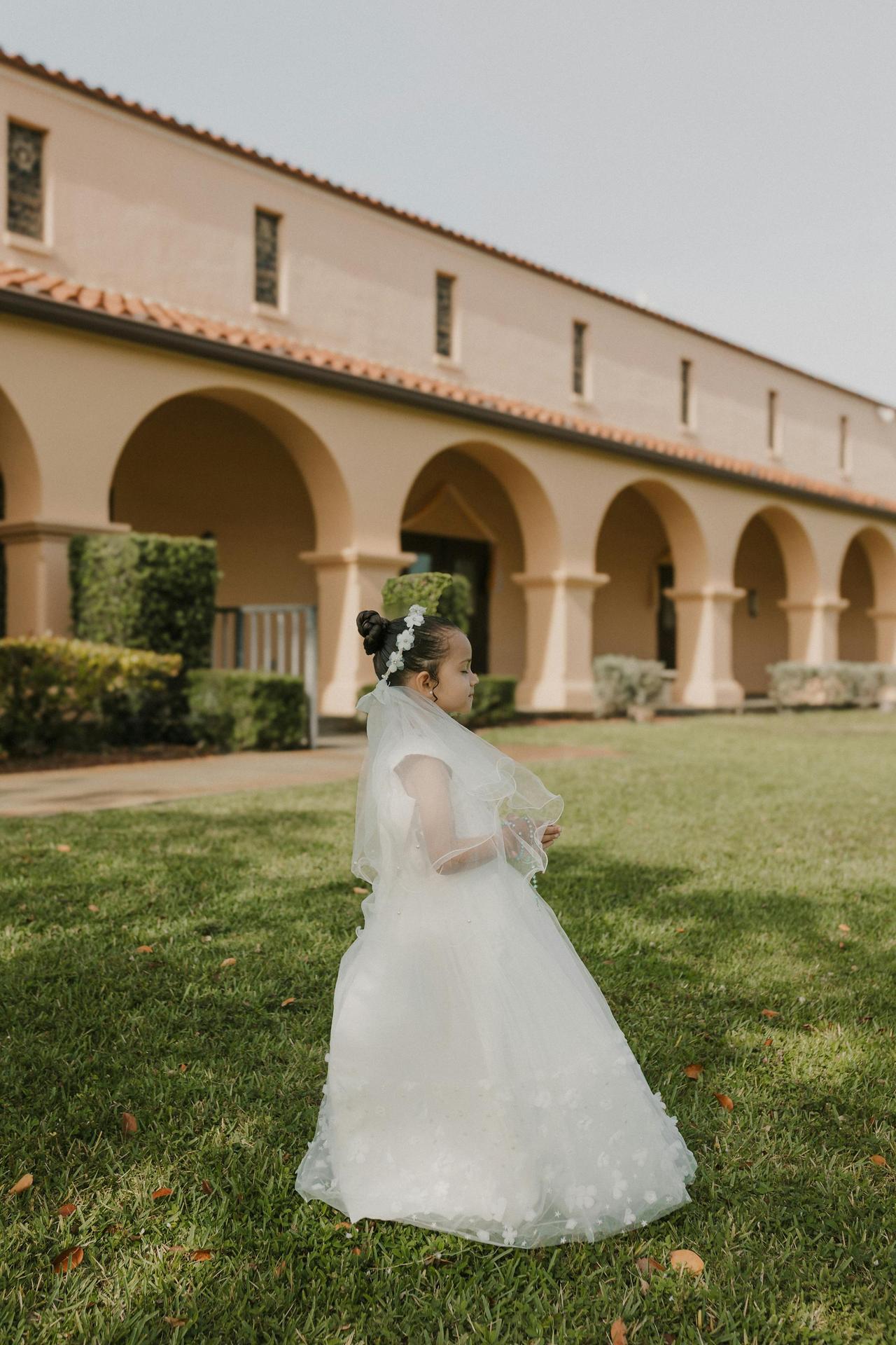 A young girl in a white communion dress stands outside a historic building with arches.