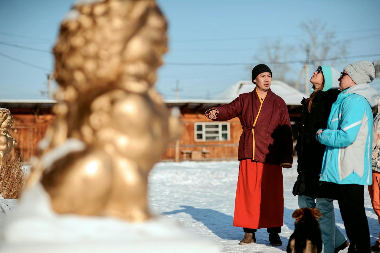 A monk in traditional attire interacts with tourists in a snowy village, with golden sculptures visible.