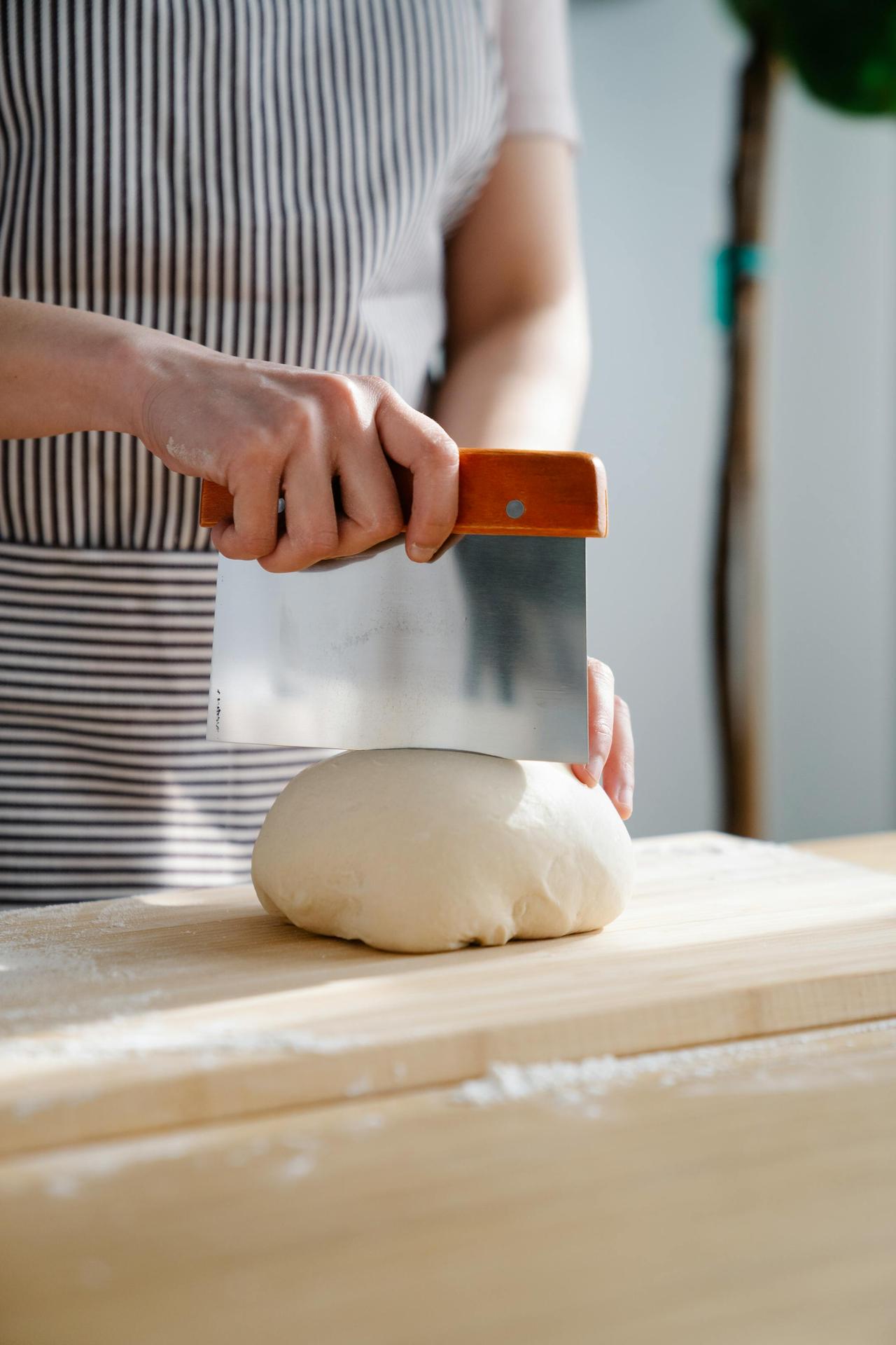 A baker uses a bench scraper to divide dough on a wooden board indoors.