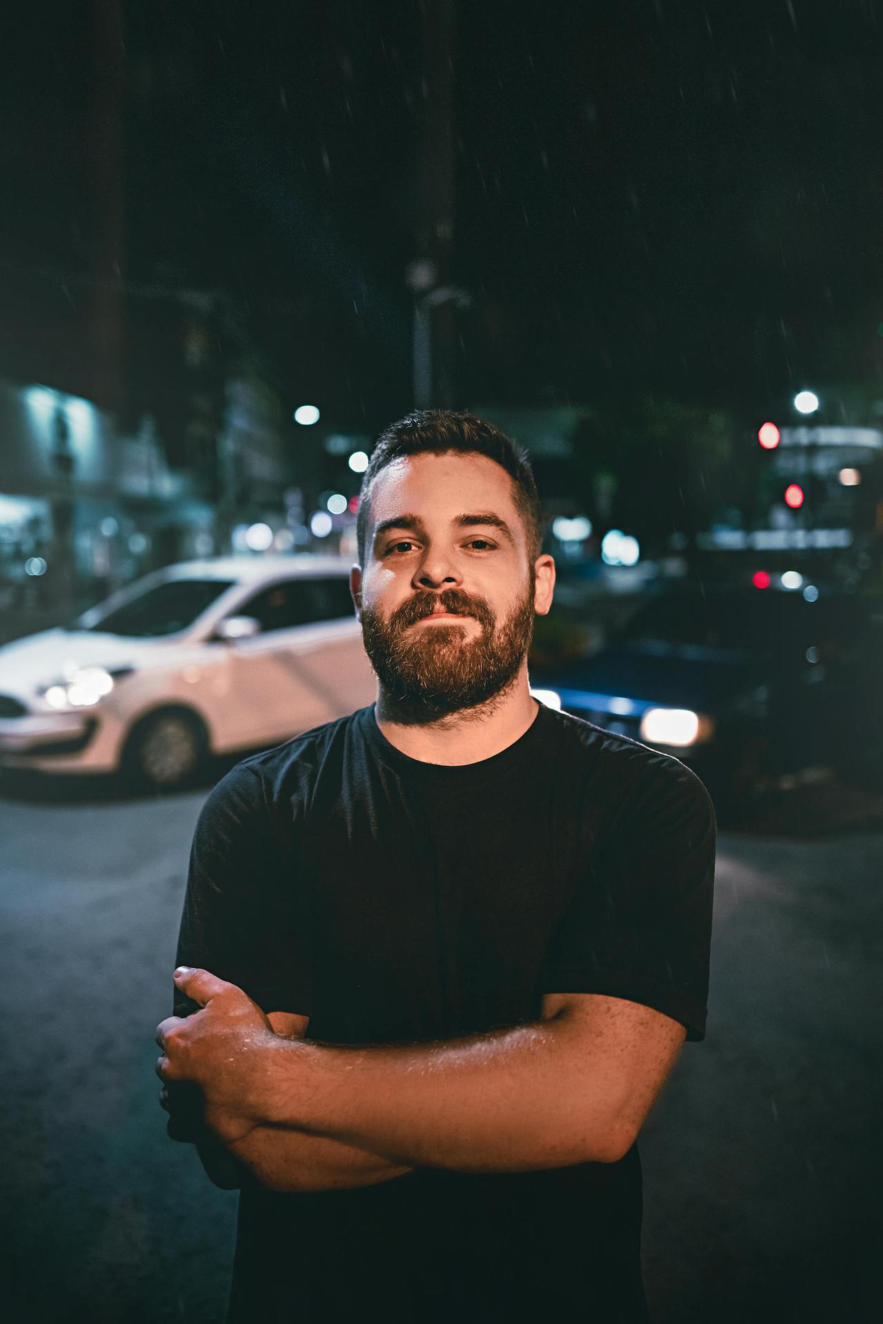 A bearded man with arms crossed stands confidently on a city street at night.