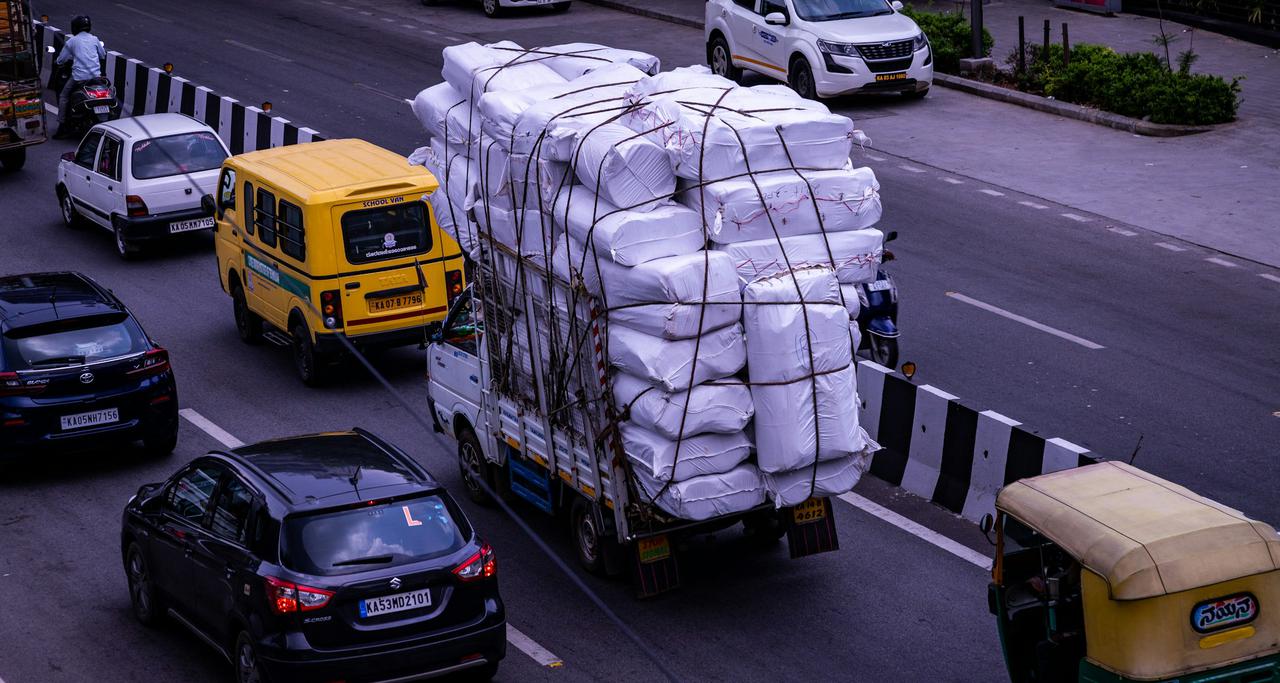 A heavily loaded truck carrying an oversized cargo of white-wrapped packages secured with ropes, navigating through traffic on a busy street in Bangalore. Surrounding vehicles include cars, a yellow school van, and an auto rickshaw, depicting the dynamic urban transport and logistics scene in the city.