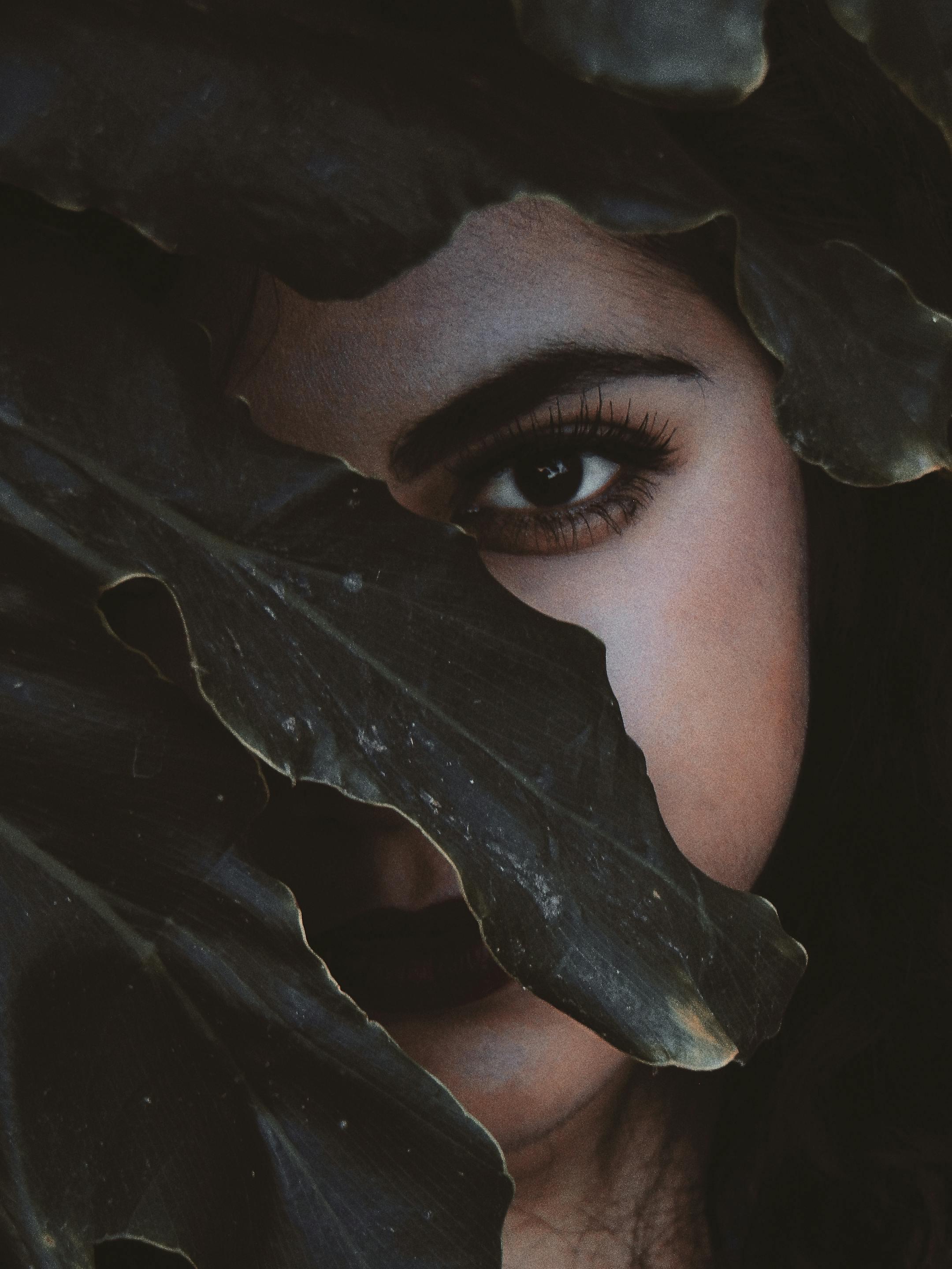 A woman's eye peers through lush dark leaves, creating a mysterious and captivating atmosphere.