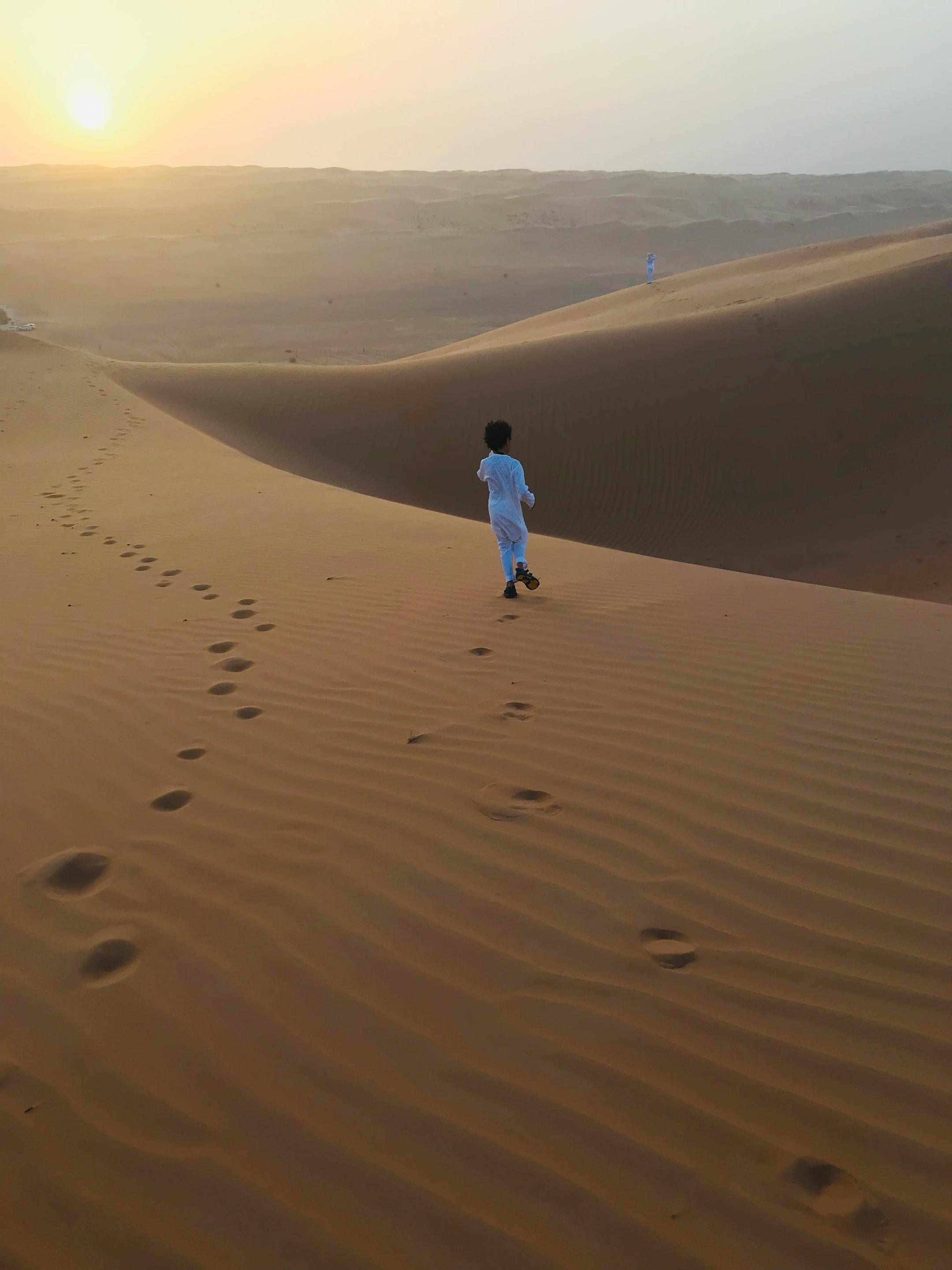 A young child in traditional attire walks on sand dunes during a warm sunset.
