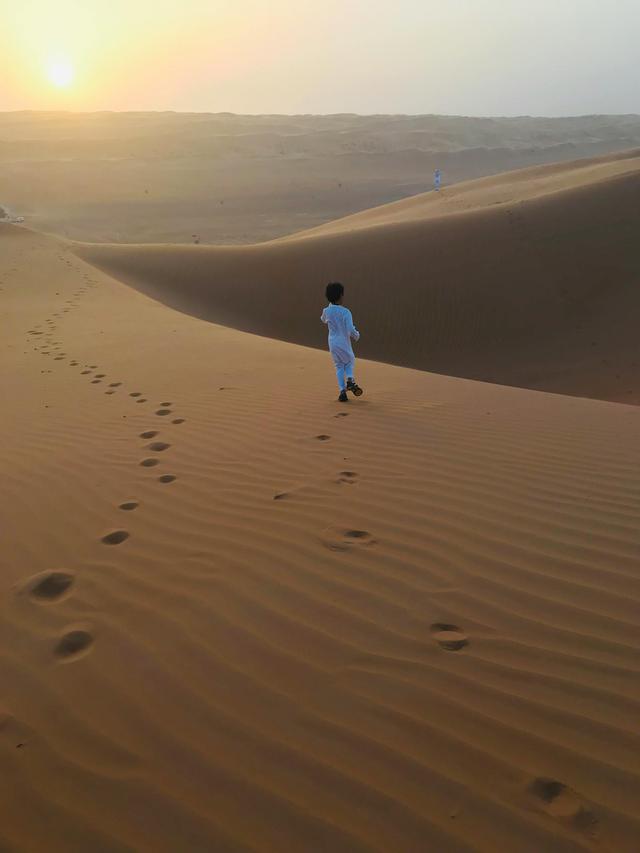 A young child in traditional attire walks on sand dunes during a warm sunset.