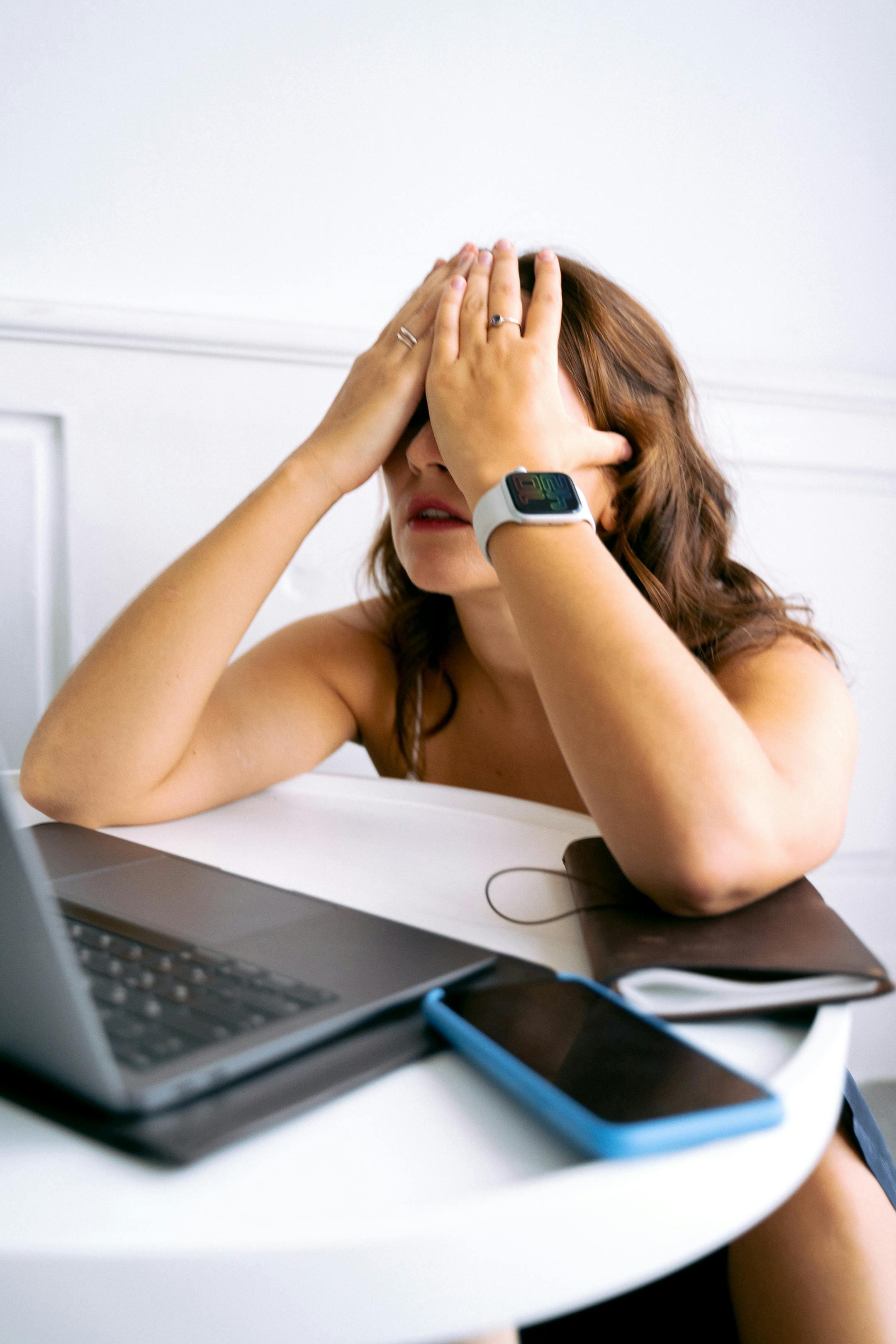 Woman showing stress or tiredness with hands on face at a desk with a laptop.