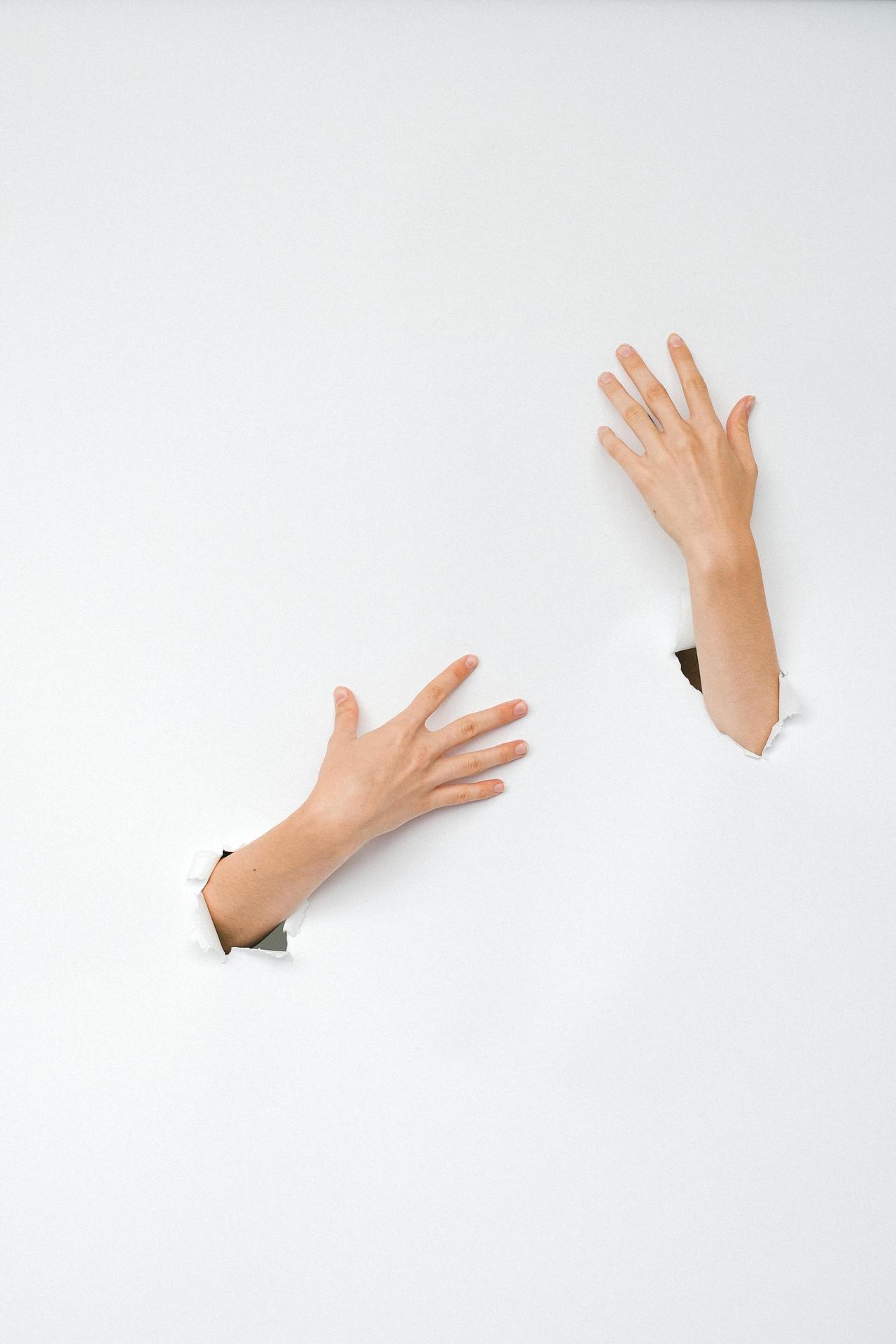 Two hands reaching through torn white paper, symbolizing emergence or escape.