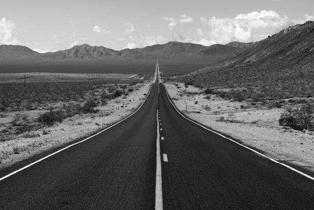 Black and white view of a long, empty highway stretching into distant desert mountains.