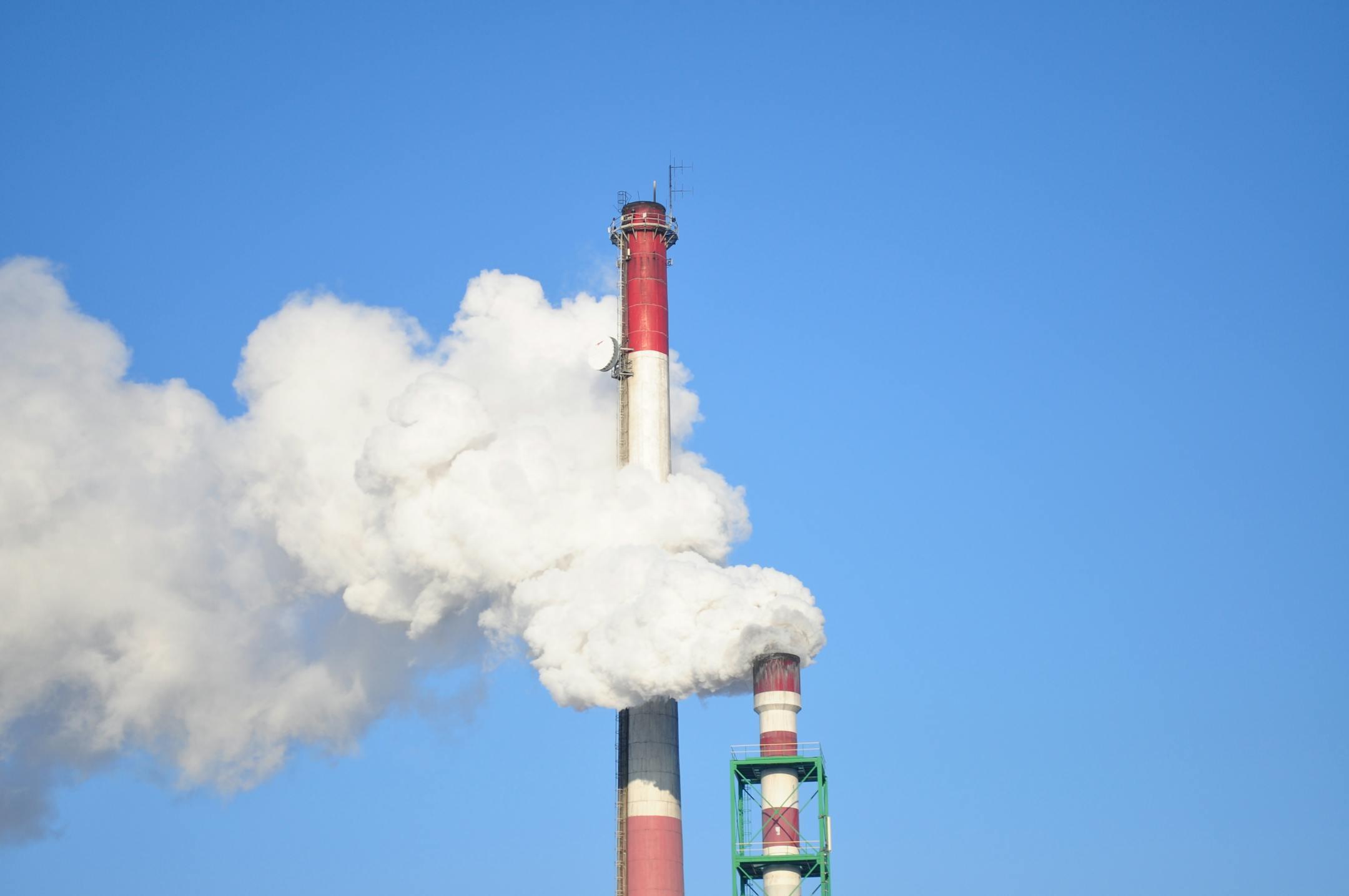 A tall industrial smokestack releasing thick white smoke against a clear blue sky.