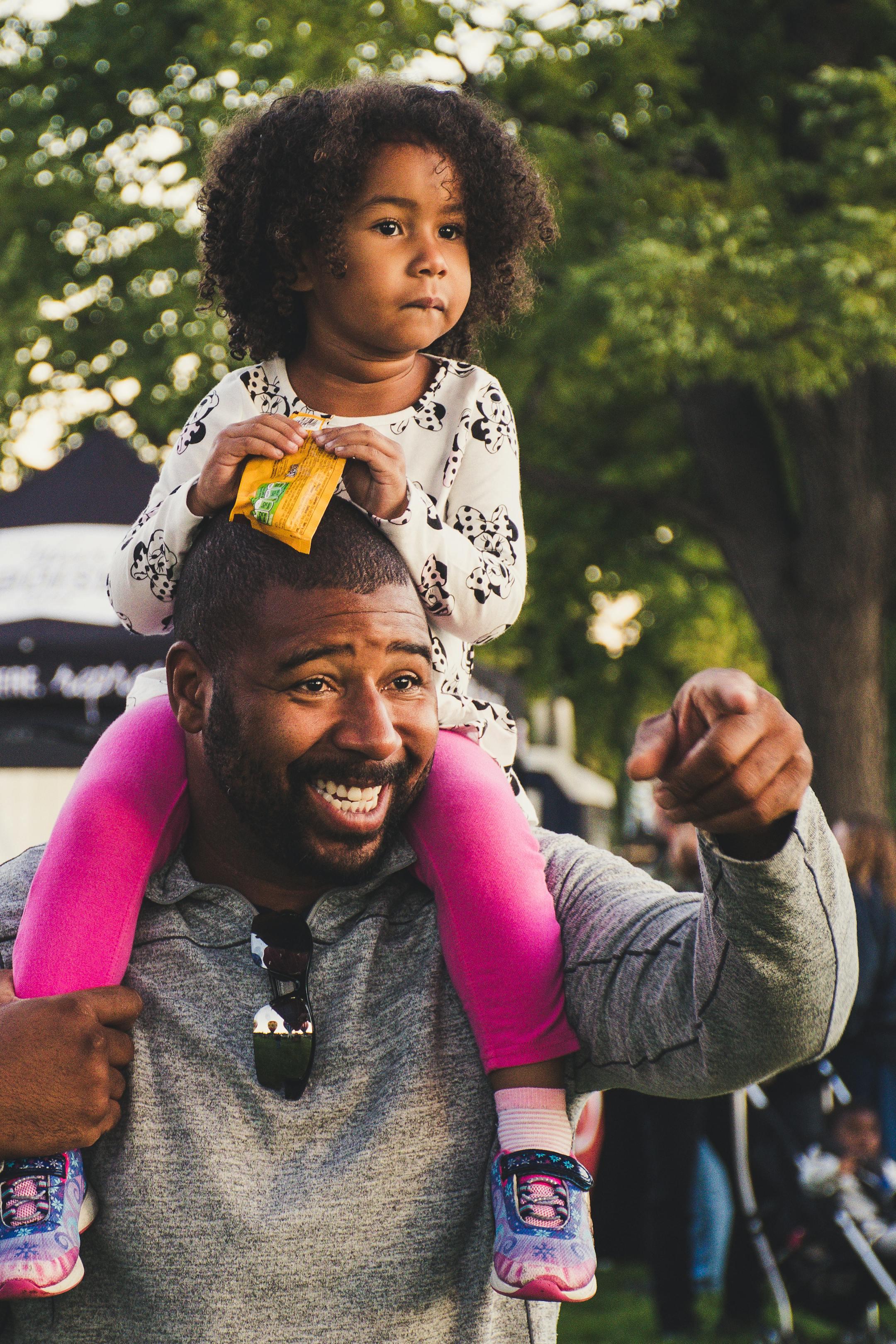 A joyful African American father with curly-haired daughter on shoulders, outdoors.
