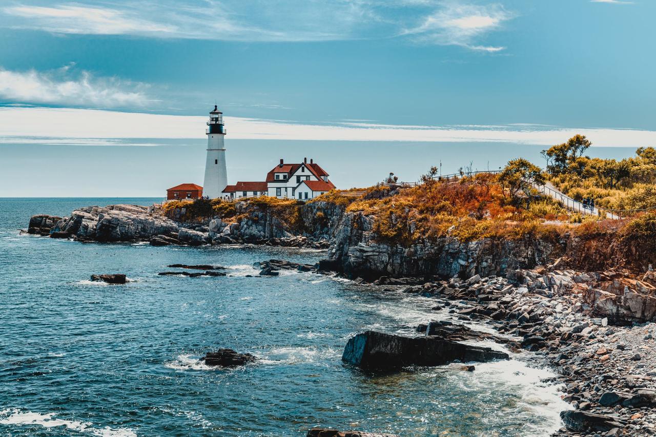 Picturesque view of wavy ocean with lighthouse and rocky formations with plants under blue sky