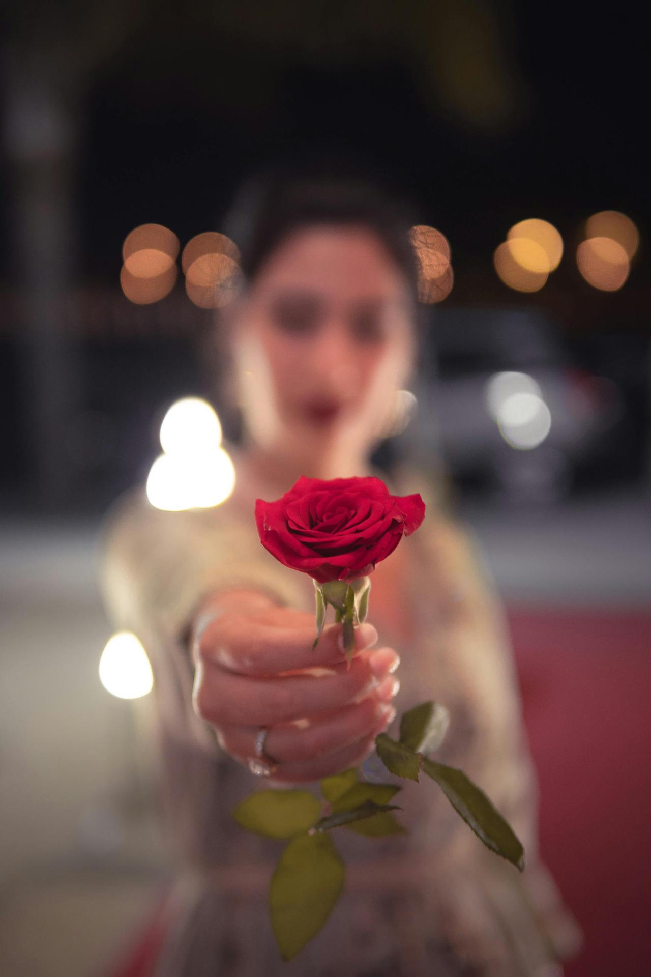 Blurry woman holding a vibrant red rose in focus against a bokeh-lit background.