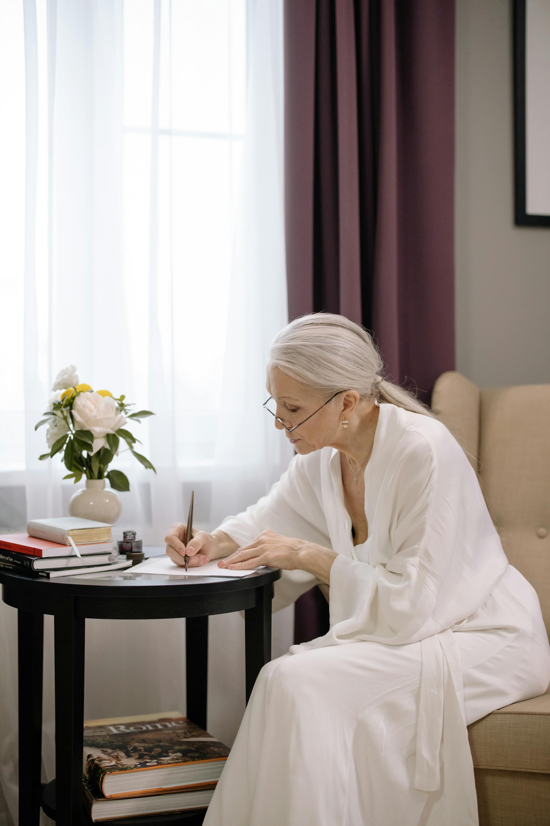 Senior woman in a white robe writing at a table beside a sunlit window, creating a serene moment.