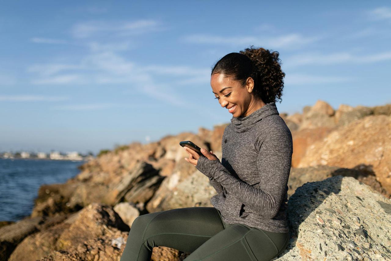 Smiling woman using smartphone on rocky shore, enjoying a sunny day.