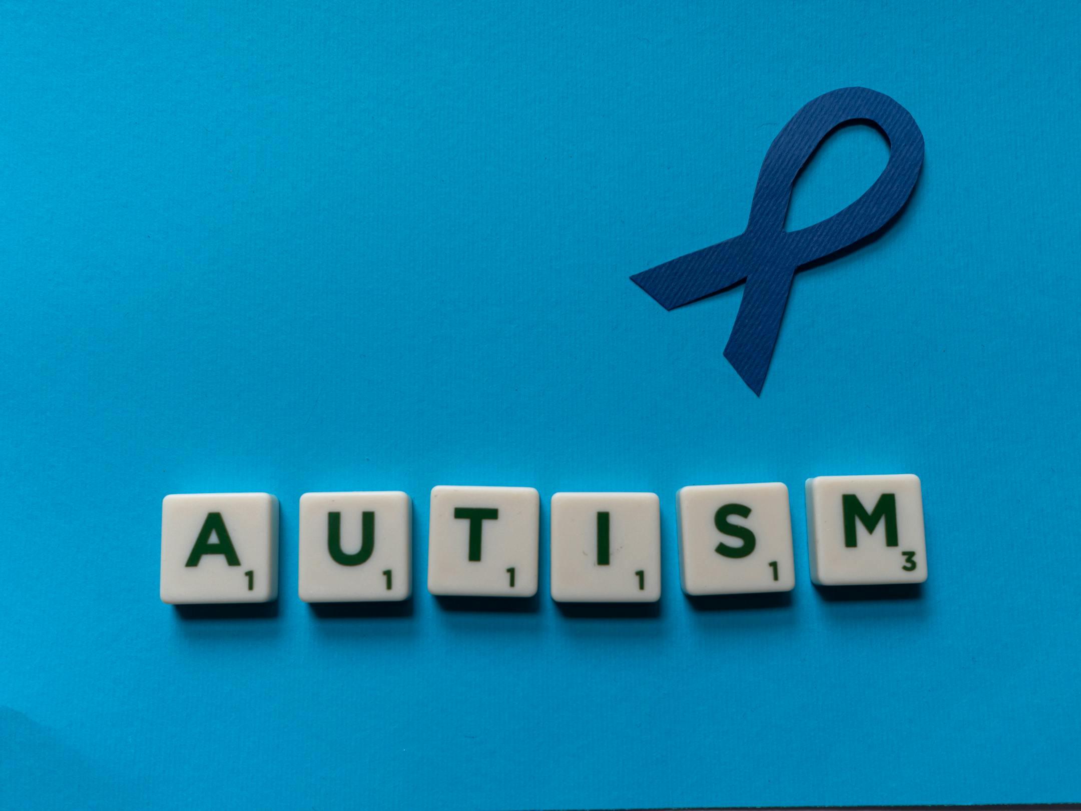 Flat lay of scrabble tiles spelling autism with a blue ribbon on a blue background.