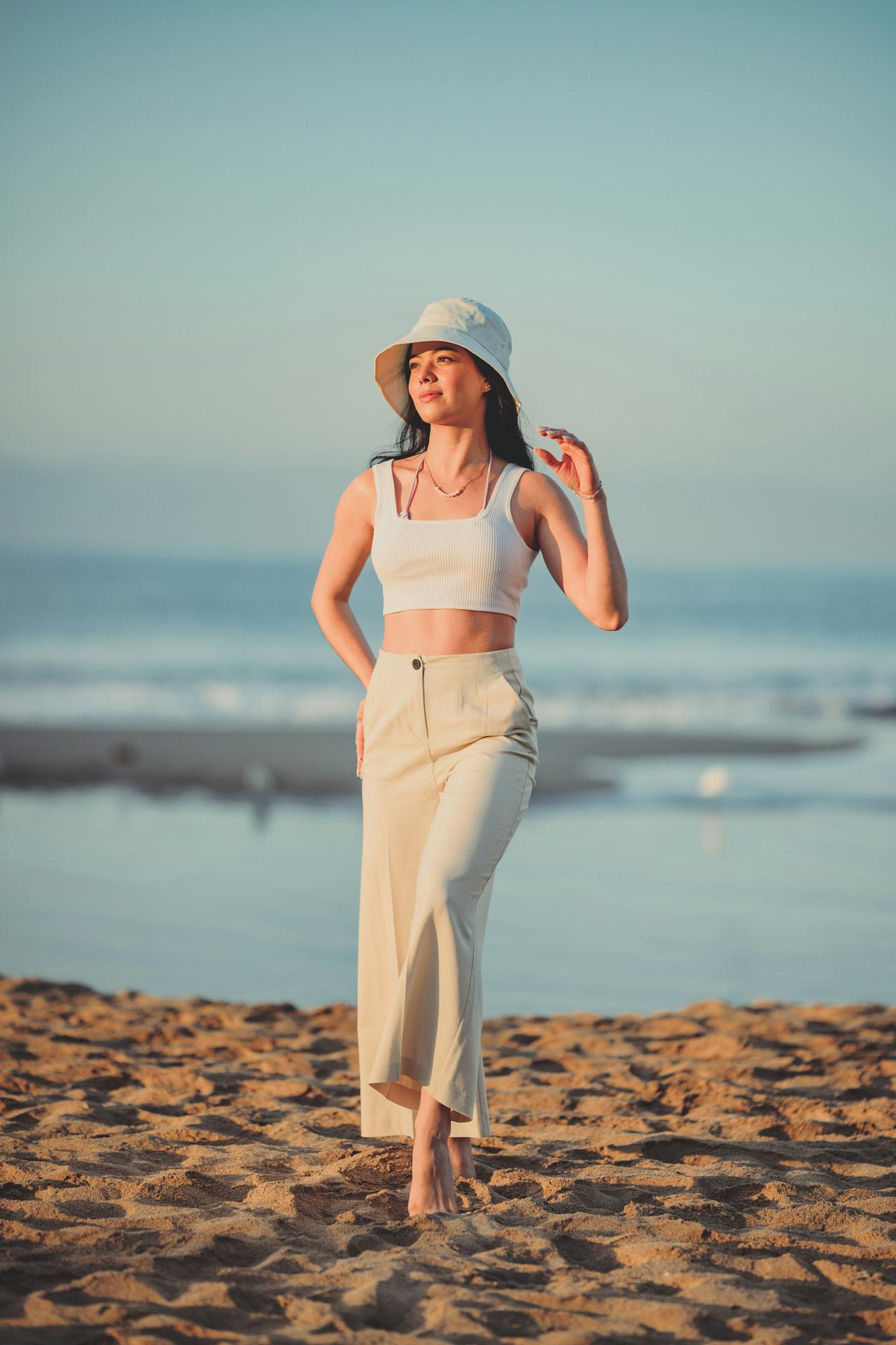 Woman modeling at the beach wearing white pants, a white hat, and a white top.