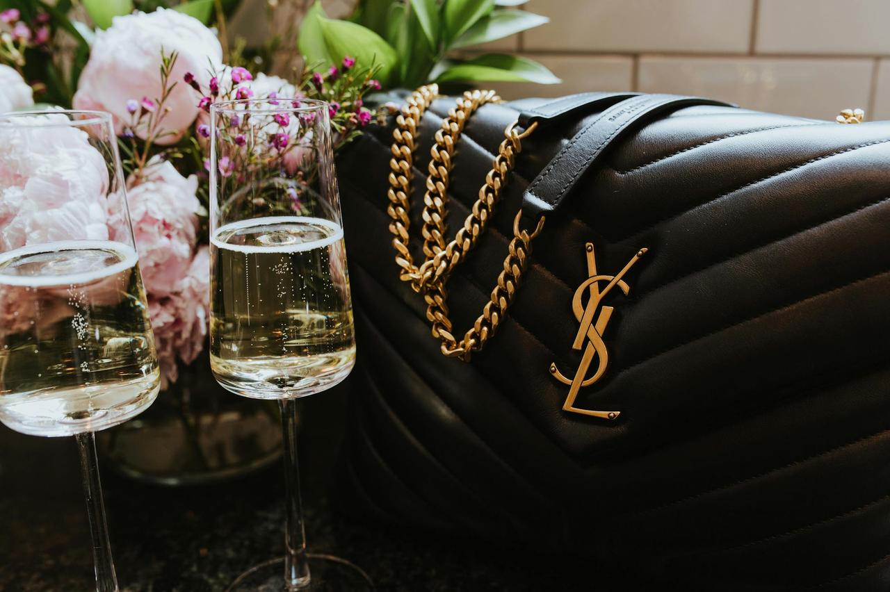 Elegant composition featuring champagne flutes and luxury designer bag amidst floral backdrop.