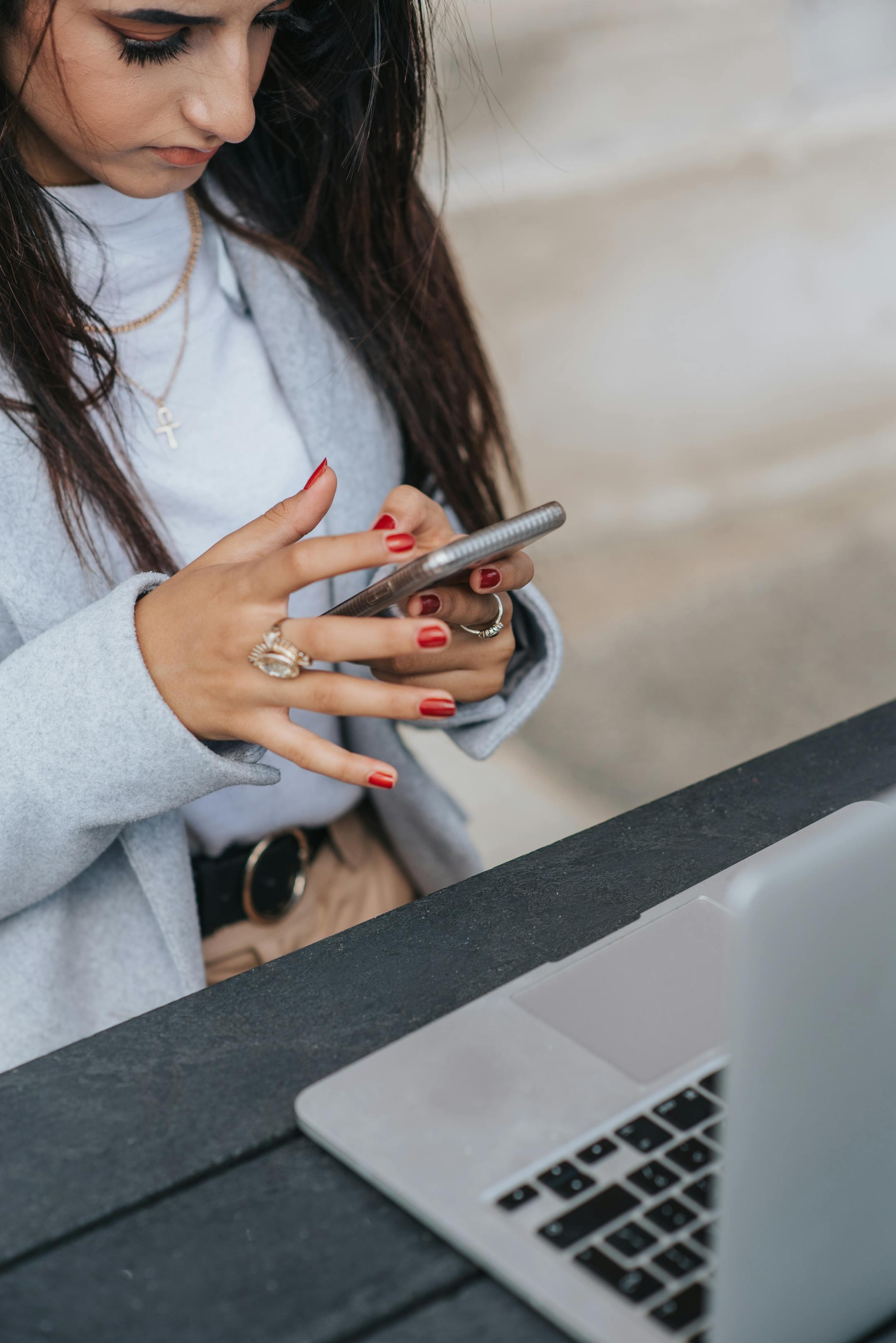 Focused businesswoman texting on smartphone while working with laptop at an outdoor cafe table.