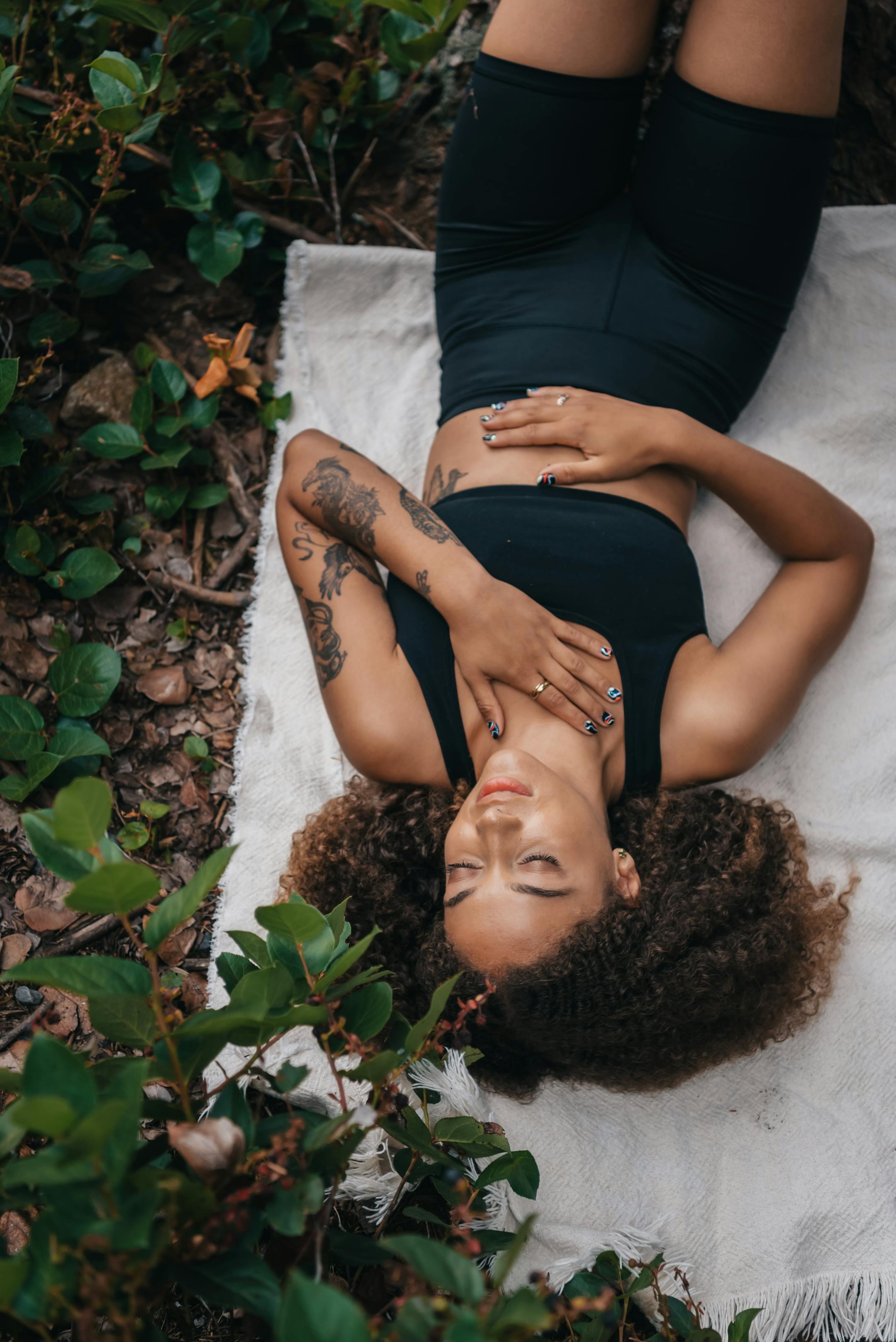A woman with tattoos and curly hair rests on a blanket outdoors, surrounded by greenery.
