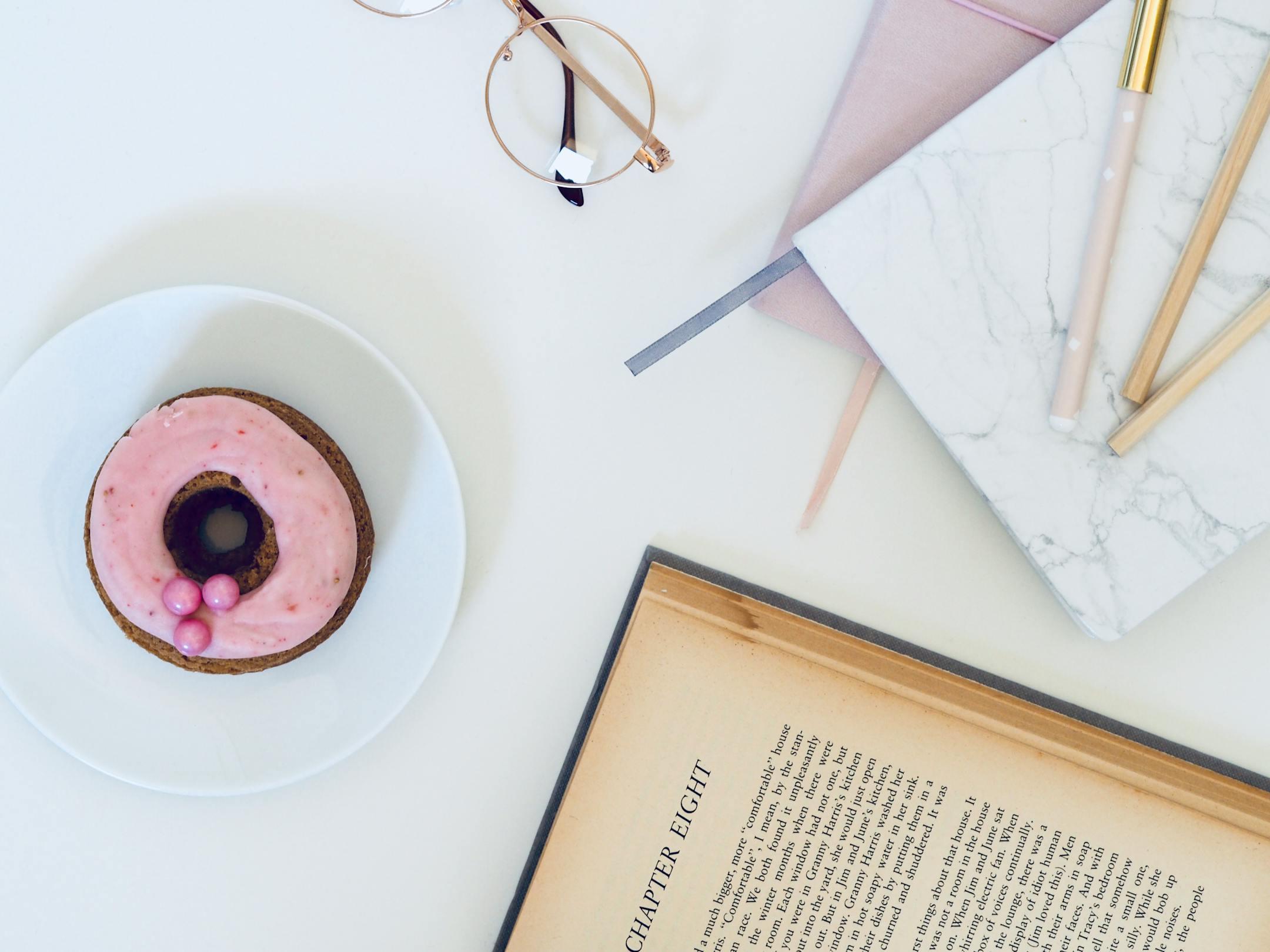 A stylish desk setup with a donut, eyeglasses, book, and notebooks. Ideal for work or study inspiration.