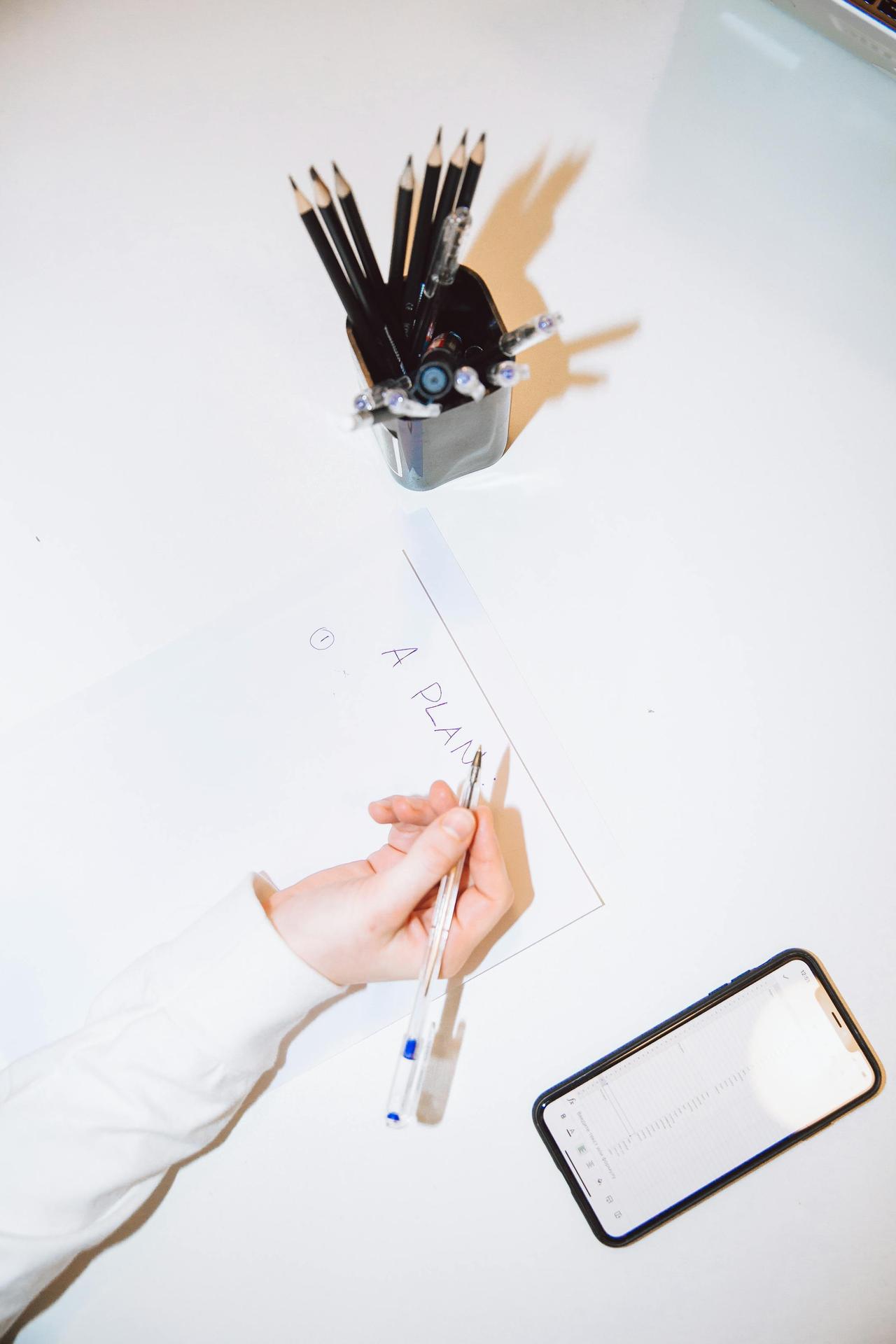 An overhead view of a woman planning on paper with a smartphone and pencils at a desk.