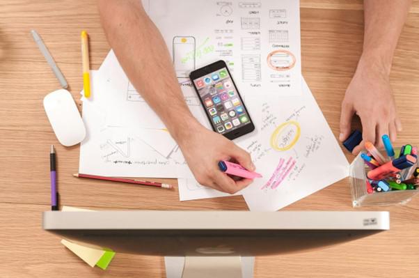A person is working at a wooden desk cluttered with papers, highlighters, pens, and a smartphone. The papers include sketches and wireframes for a mobile app design. One hand is holding a pink highlighter, marking notes on a sheet, while the other hand rests near a container filled with colorful markers. An iMac computer and a wireless mouse are also visible on the desk.
