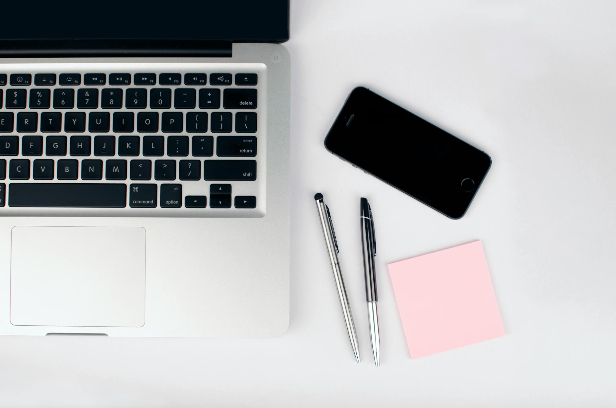 A top view of a modern workspace featuring a laptop, smartphone, pens, and a pink sticky note on a white desk.