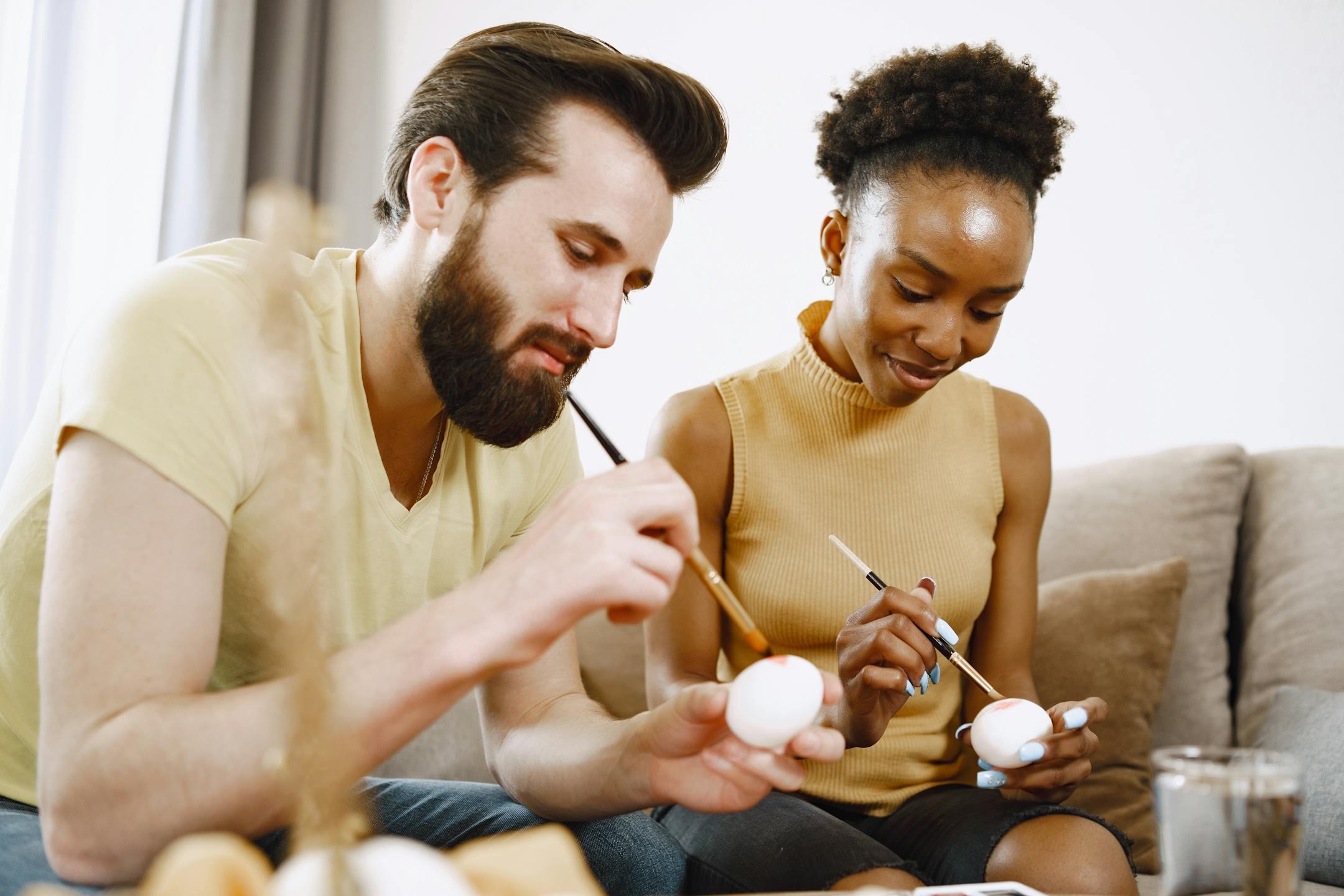 A multiracial couple painting eggs indoors, celebrating Easter, and enjoying togetherness.