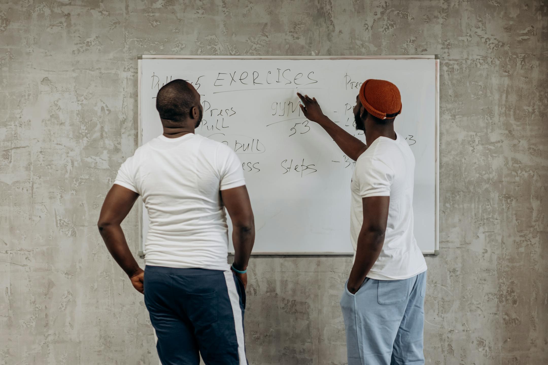 Two adult men planning their gym exercises on a whiteboard in a modern setting, emphasizing fitness and health.
