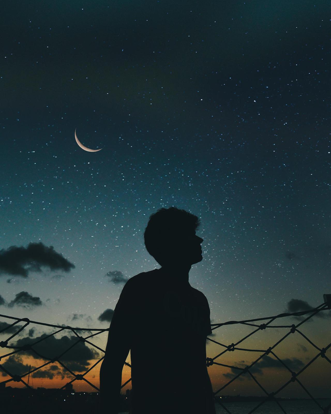 A lone silhouette stands beneath a crescent moon and starry sky, evoking a sense of solitude and wonder.