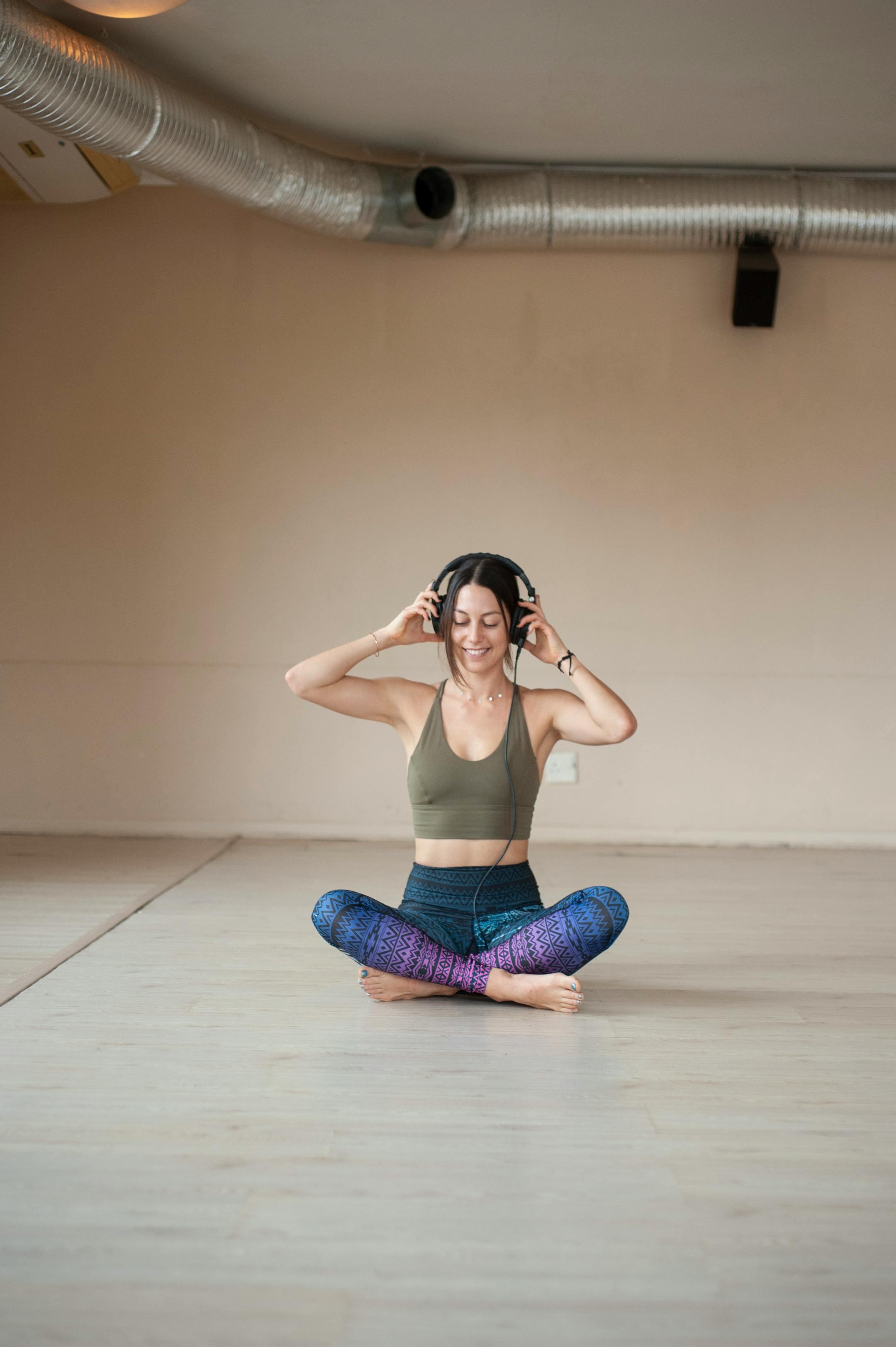 A smiling woman in activewear sits cross-legged, listening to music in a yoga studio.