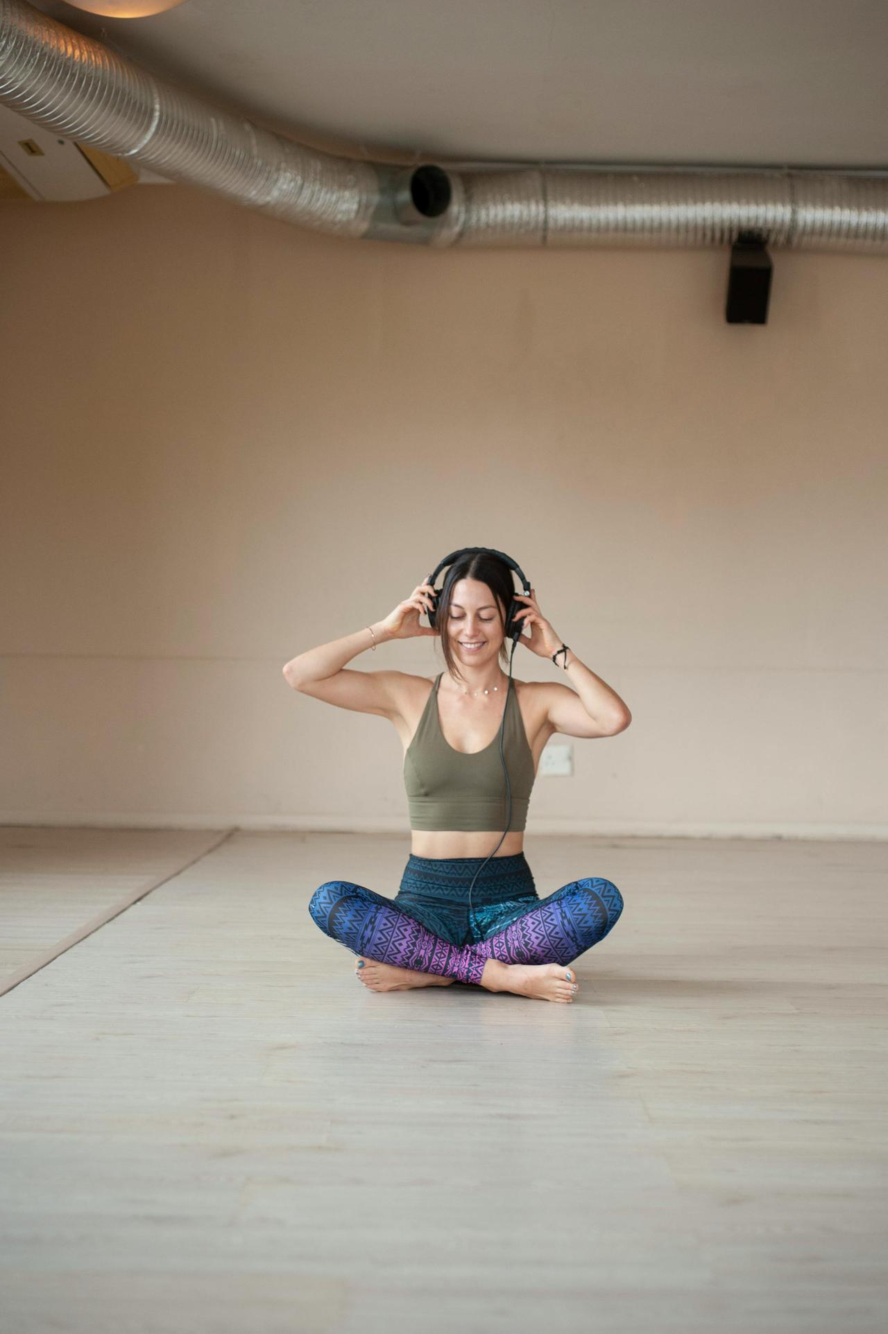 A smiling woman in activewear sits cross-legged, listening to music in a yoga studio.