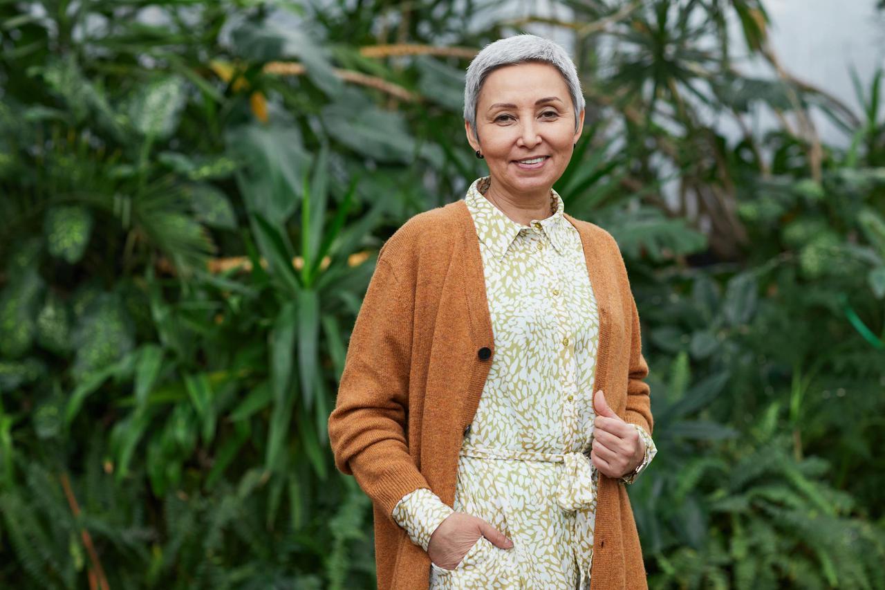 Portrait of a joyful senior woman in a stylish cardigan and dress, smiling in a vibrant greenhouse setting.
