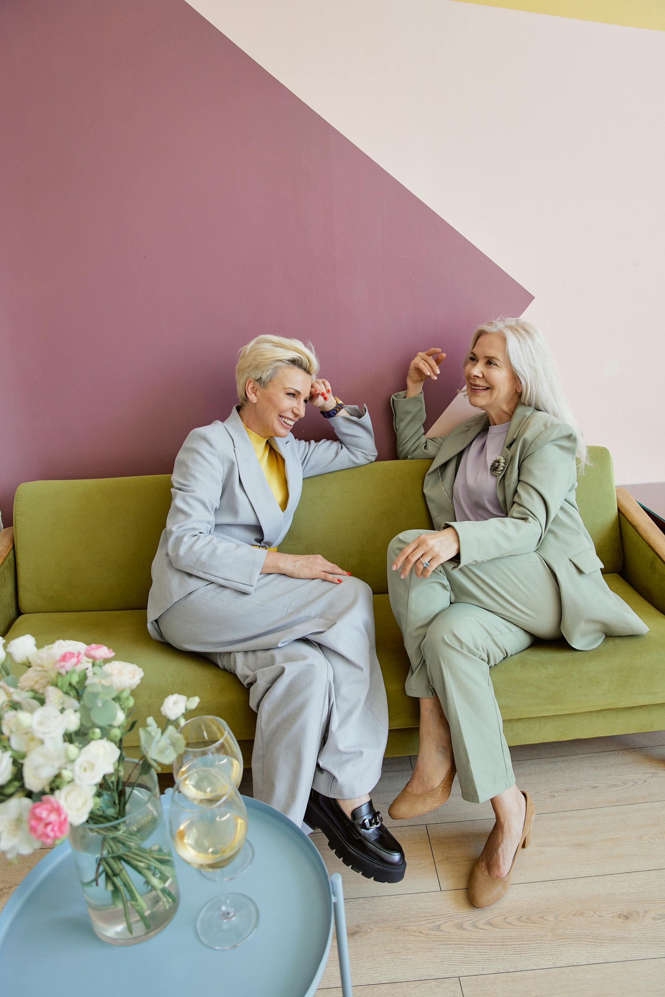 Two stylish senior women in suits smiling and chatting on a modern sofa, enjoying each other's company indoors.