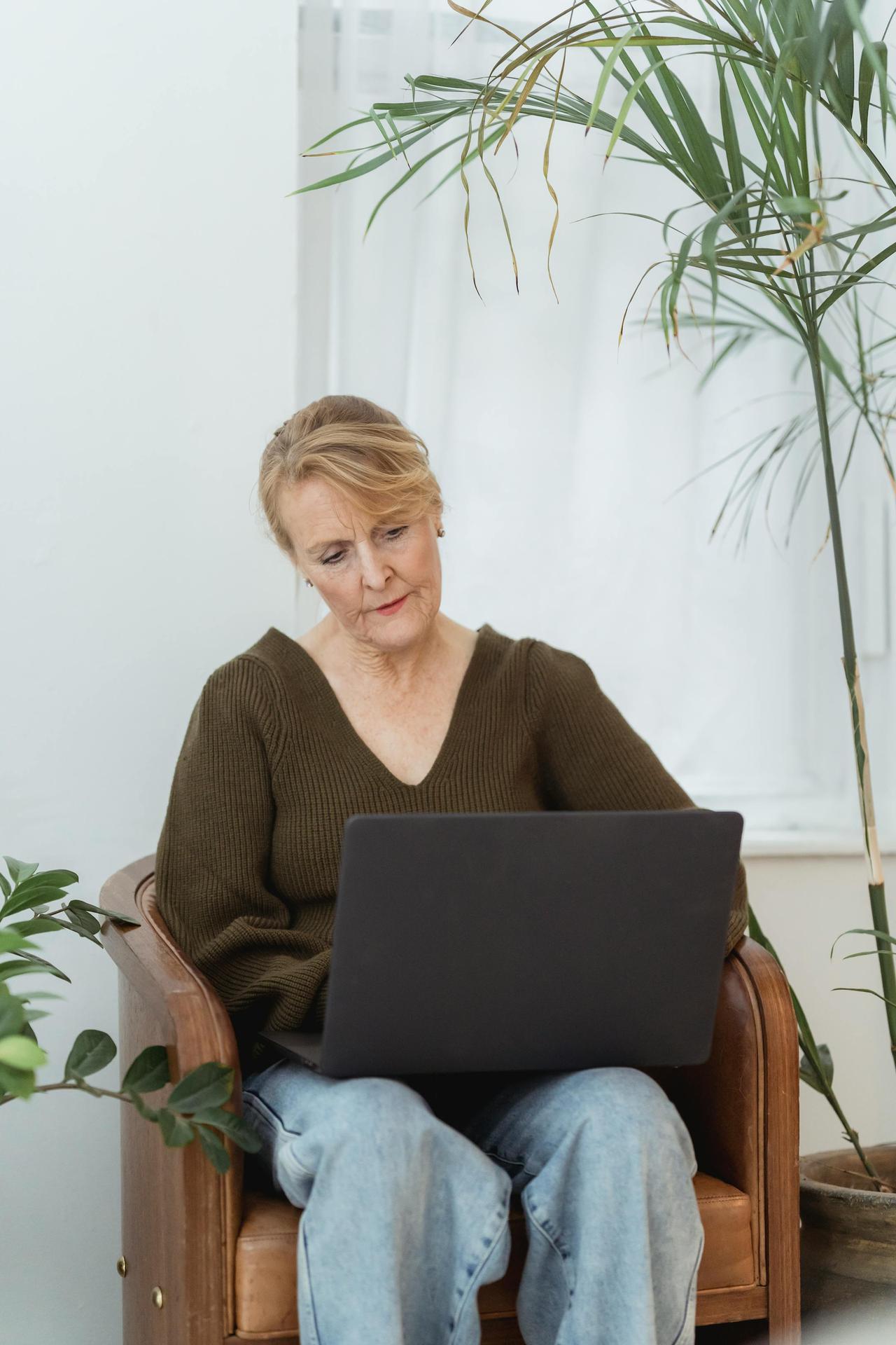 A senior woman sits attentively with a laptop, surrounded by plants, in a cozy living room setting.