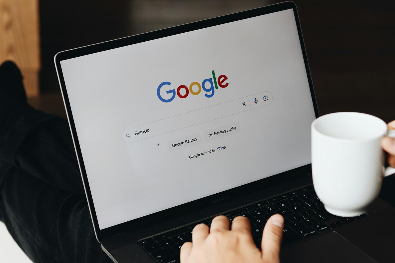 A person is using a laptop to search for "SumUp" on Google while holding a white coffee mug. The Google search page is displayed on the screen, and the user is preparing to click "Google Search."