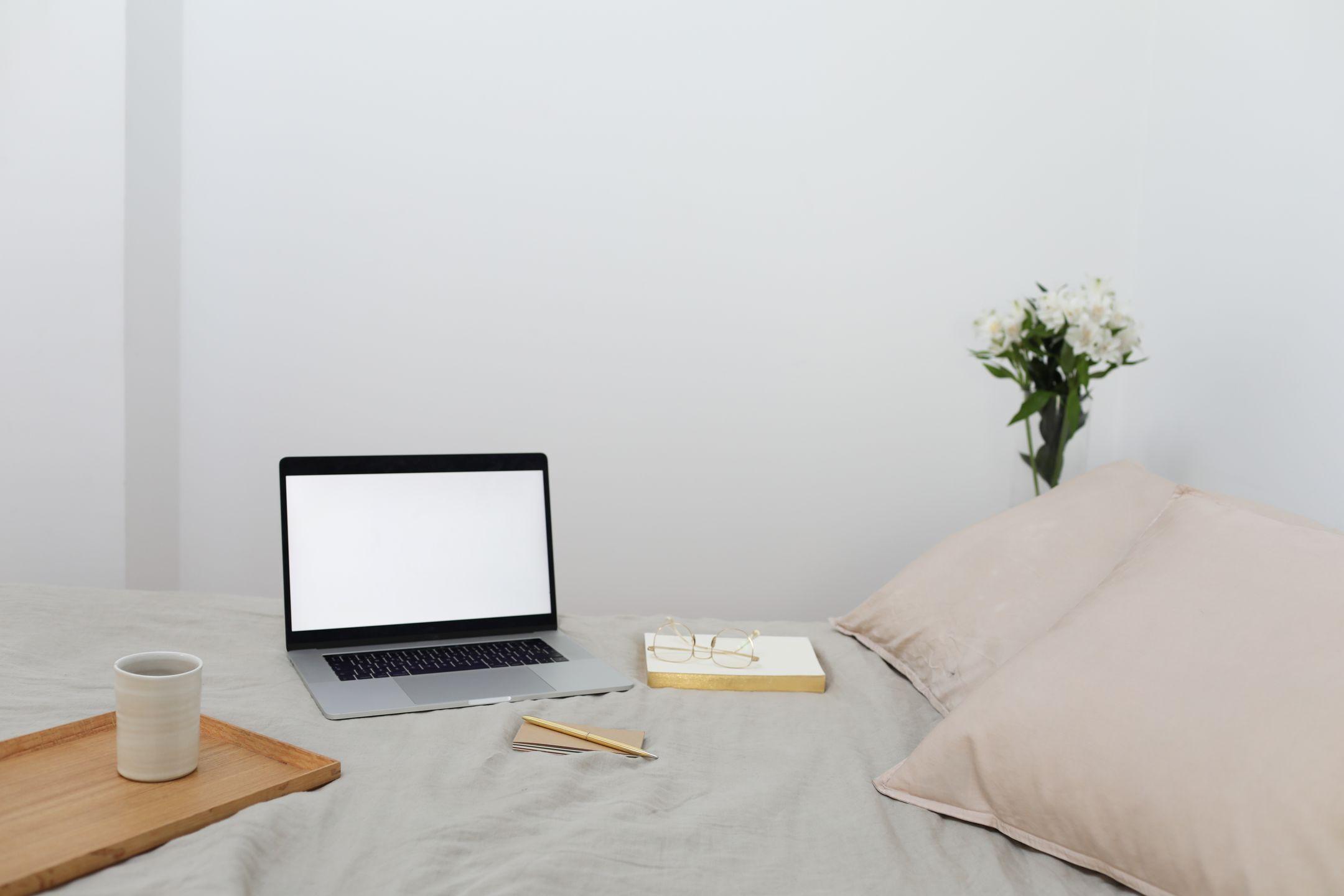 Wooden tray with coffee cup and modern netbook placed on comfy bed with textbook near soft pillows during distance lesson