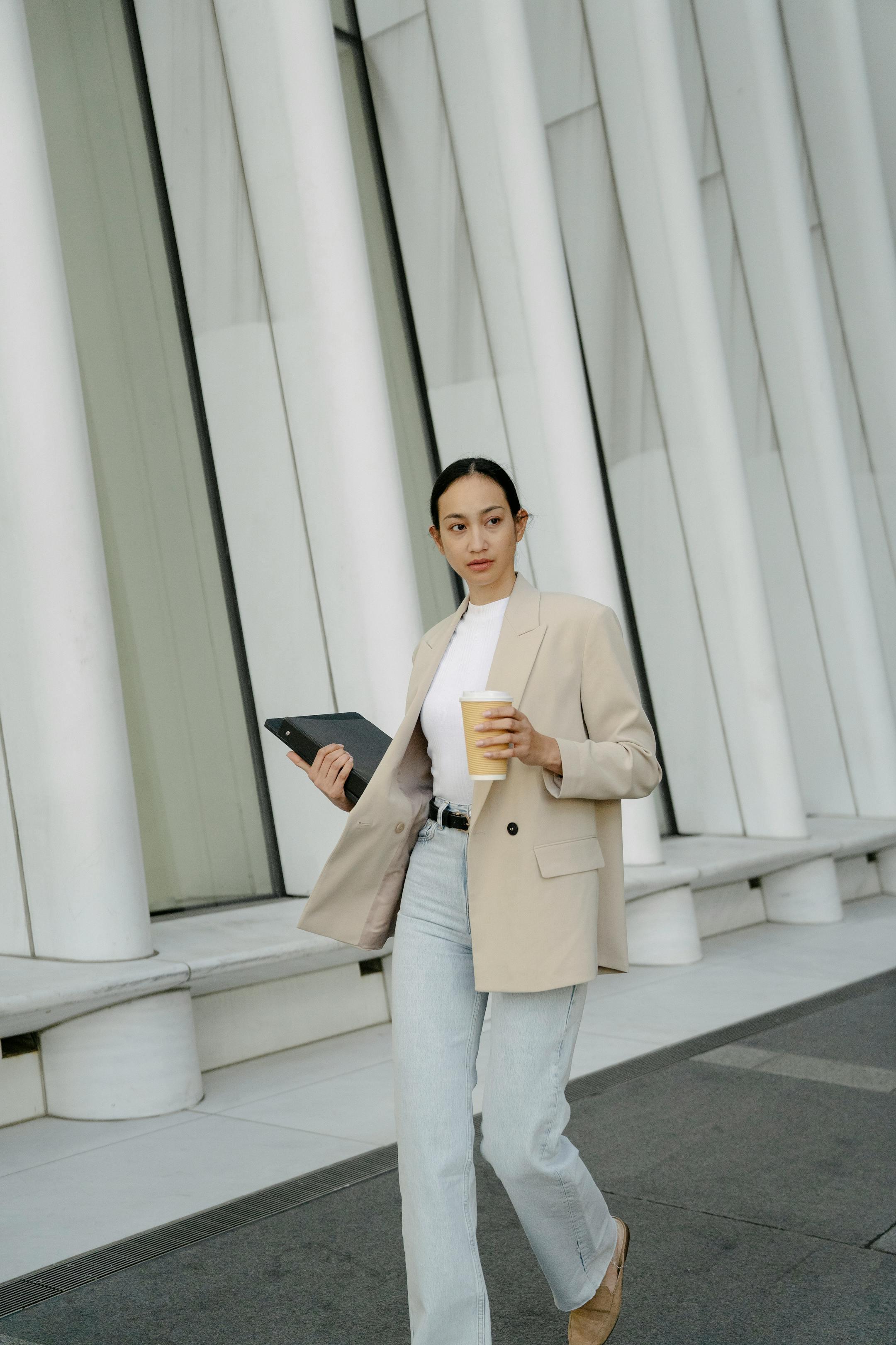 Stylish young woman walks confidently with coffee and laptop in urban setting.